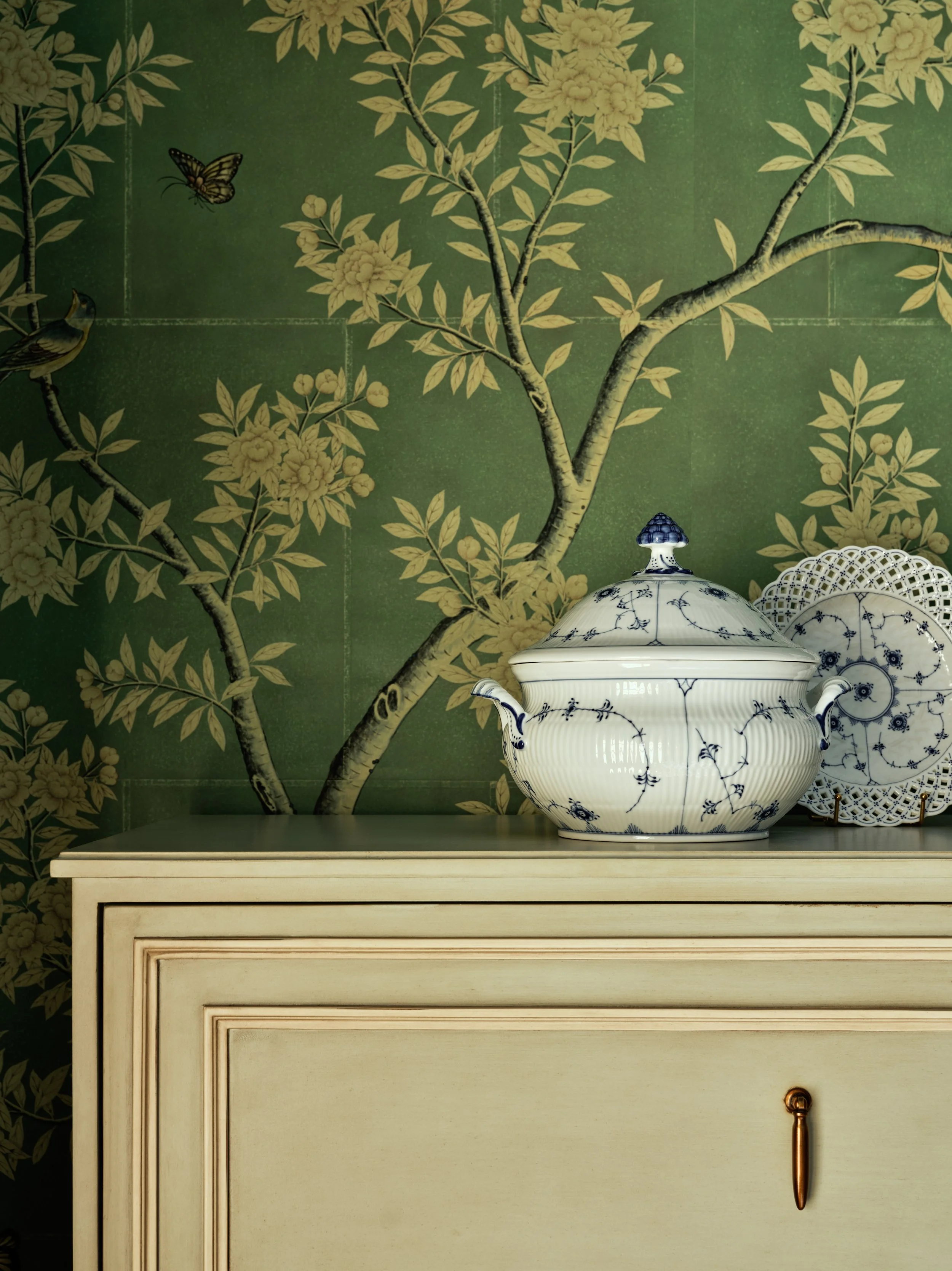 Decorative ceramic tureen with a floral pattern and a lid, and matching plate, placed on a cream-colored cabinet against a green botanical wallpaper featuring flowering trees and birds.