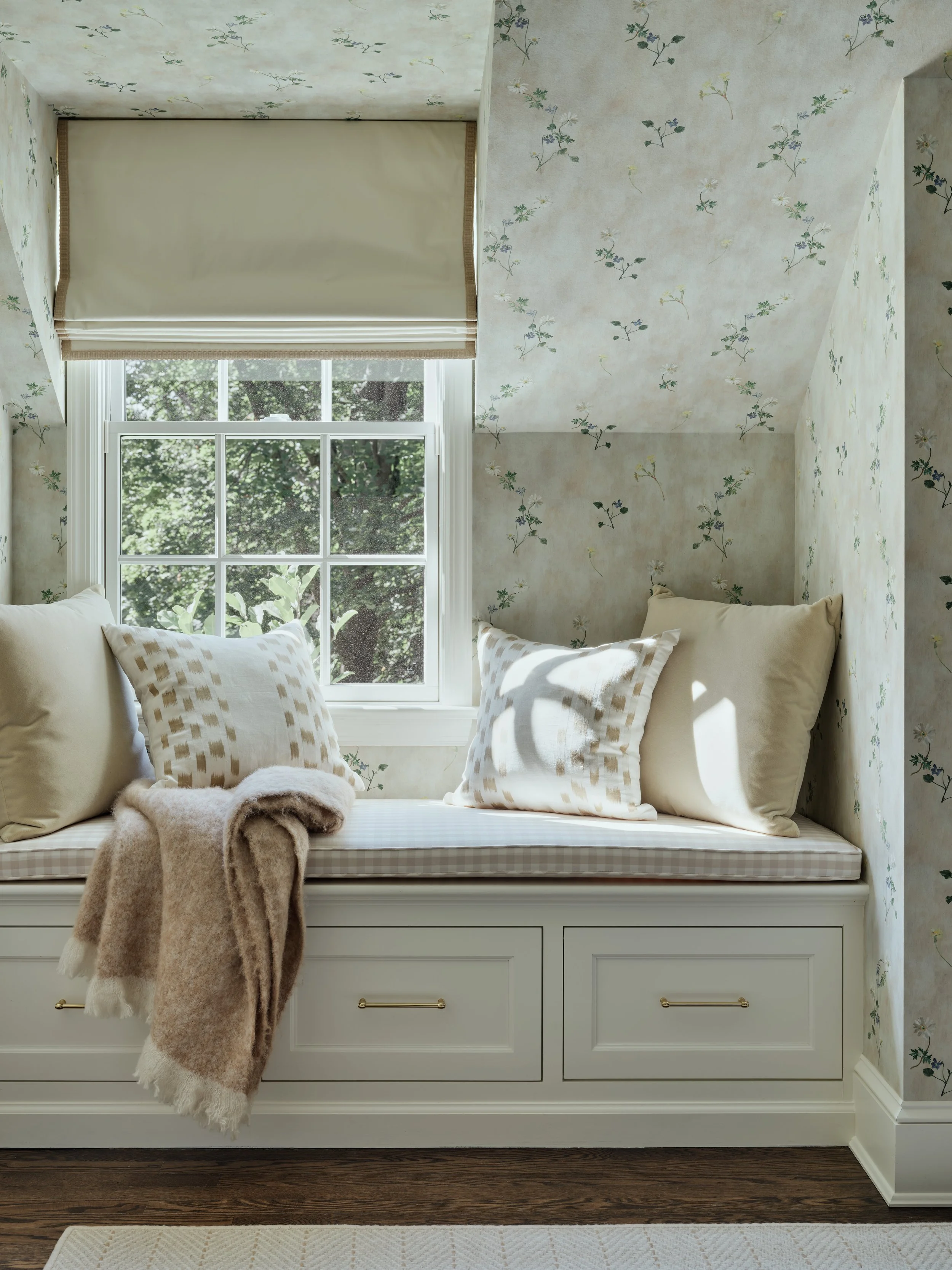 Cozy window seat with cream and beige cushions and a beige blanket, next to a window with a view of greenery, in a room with floral wallpaper.