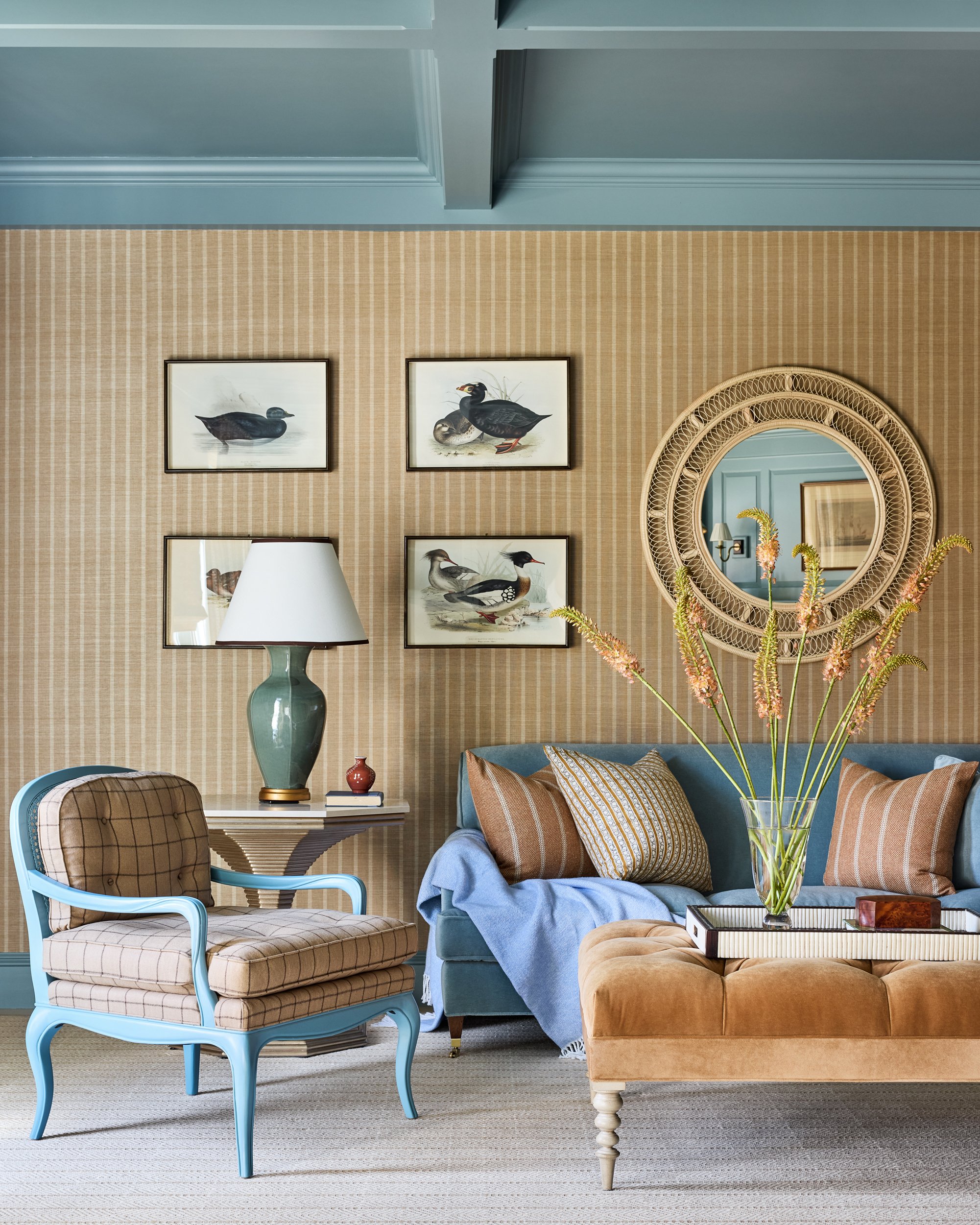 Living room with blue sofa, plaid armchair, beige ottoman, table lamp, vase with pink flowers, framed bird artwork, round mirror, striped pillows, and a blue and beige color scheme.