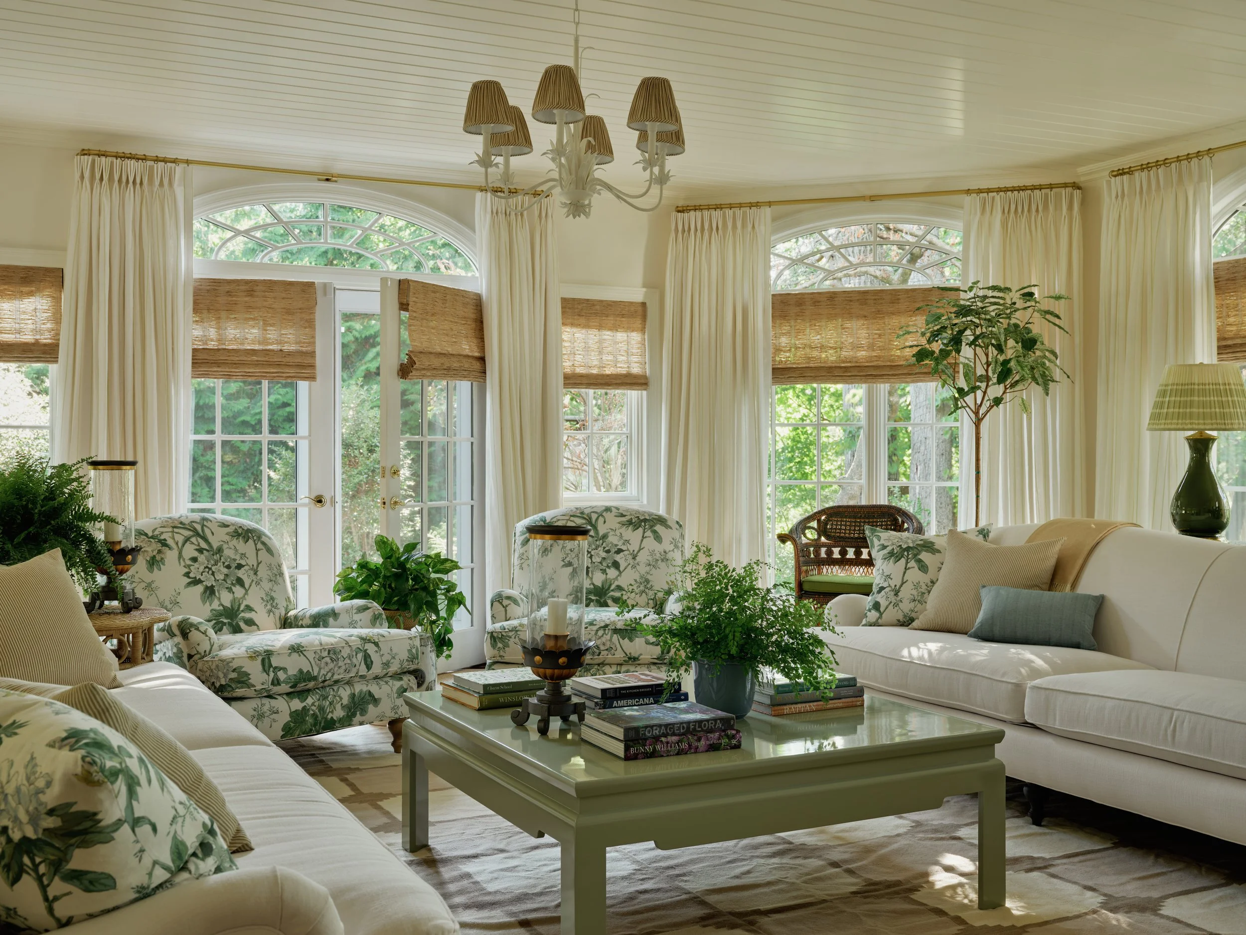 A bright, airy living room with large windows, white curtains, and woven shades. There are white and floral patterned sofas with pillows, a coffee table with books and plants, and various lamps. Greenery is visible outside.