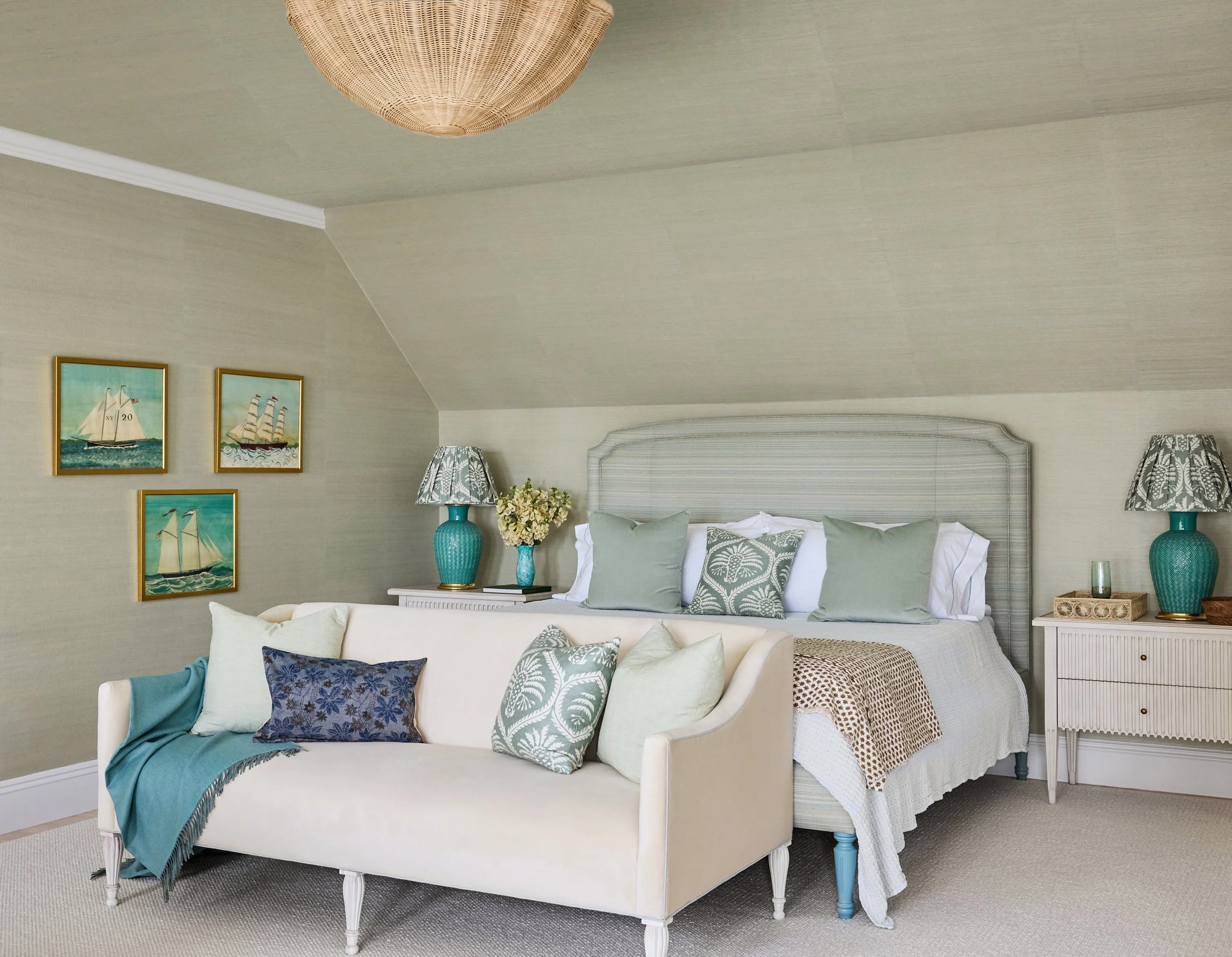 A bedroom with a bed and a sitting area, decorated in a coastal theme with ocean artwork, teal lamps, and patterned throw pillows.