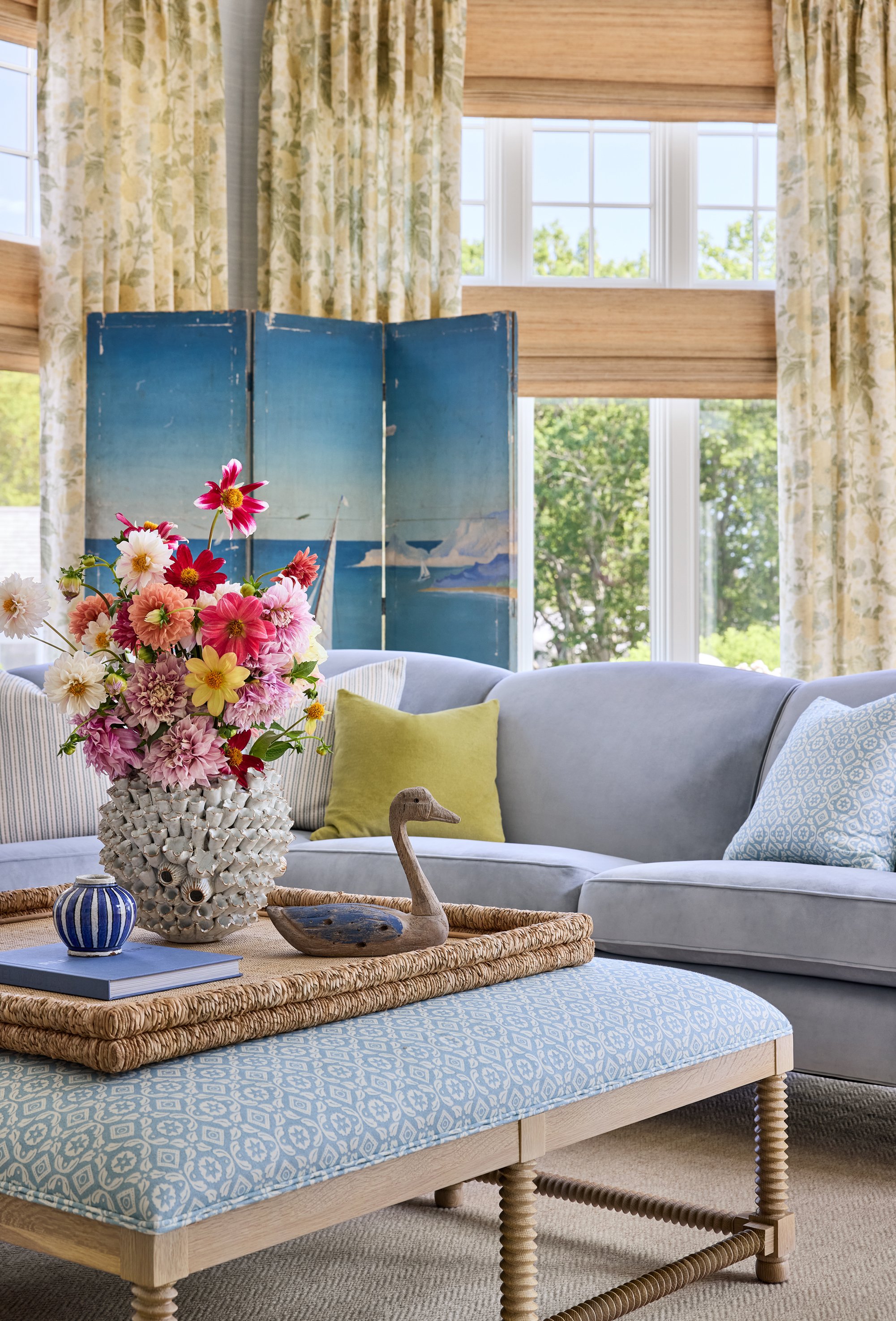 Living room with a light blue sectional sofa, decorative pillows, a rattan coffee table with a blue and white patterned cushion, a large floral arrangement in a shell-shaped vase, a wooden swan figurine, and a blue folding screen with a painted lands