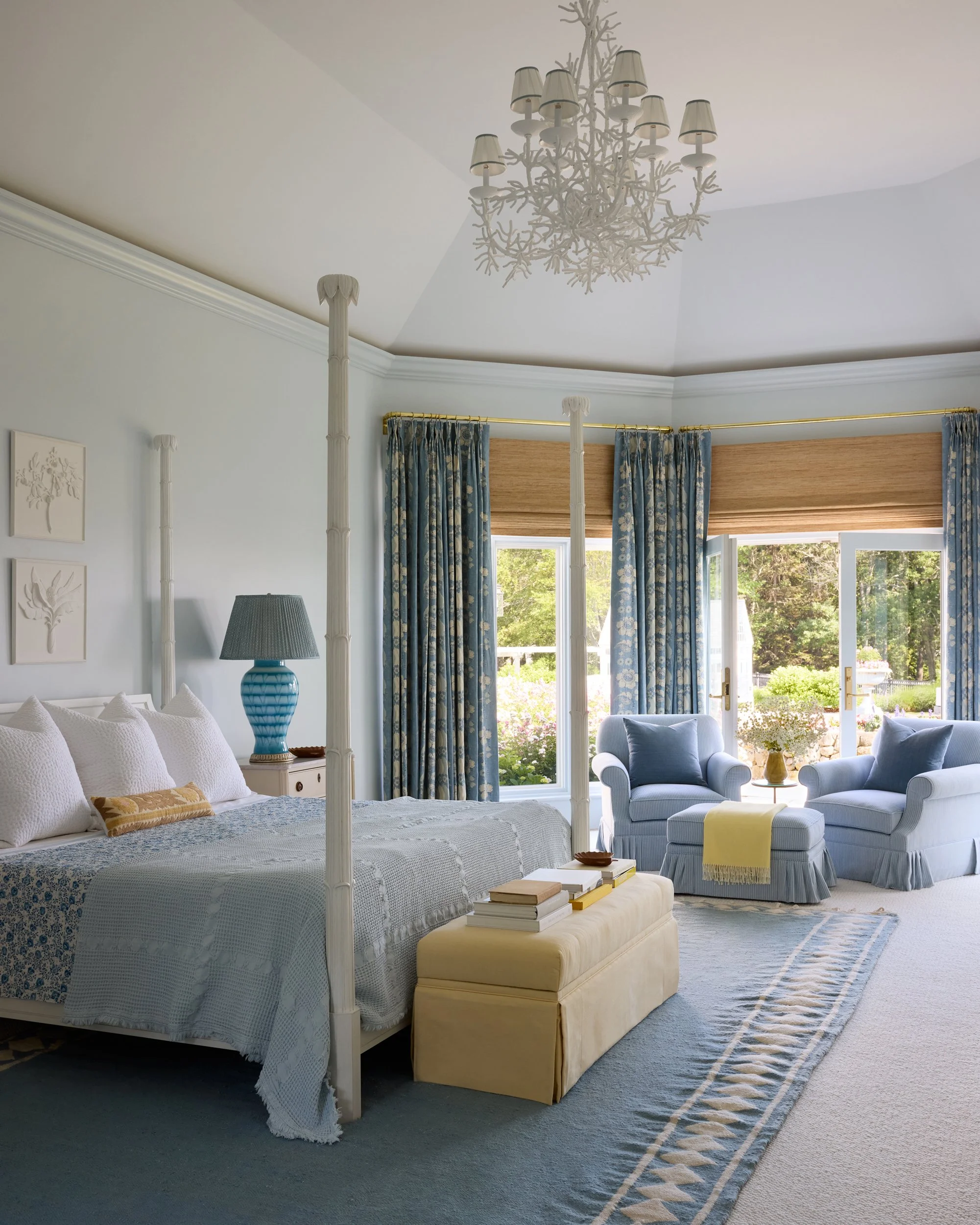 A bedroom with a four-poster bed, a blue and white color scheme, a chandelier, a blue table lamp, and two armchairs near large windows with floral curtains and views of a garden outside.