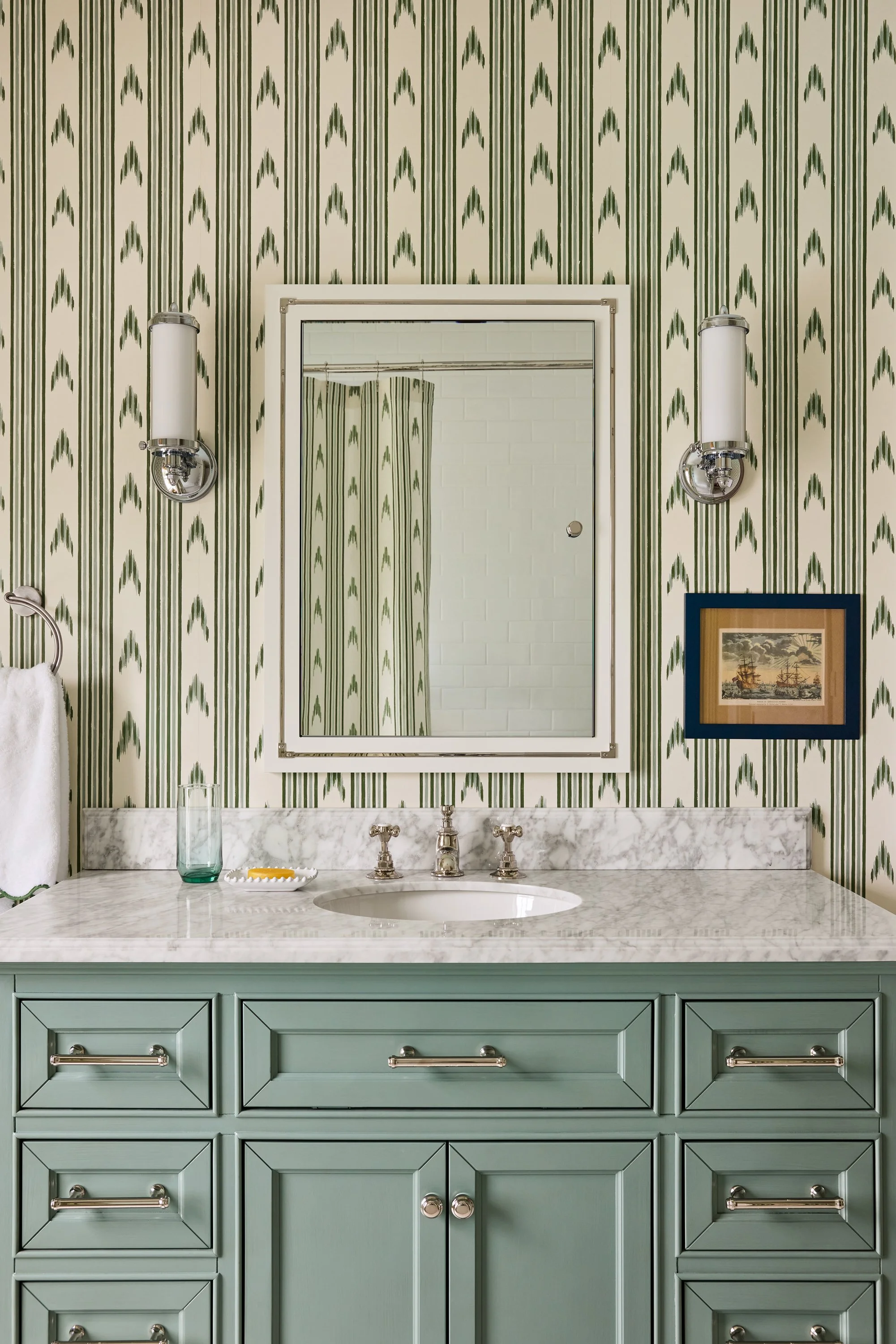 A bathroom vanity with a green cabinet and a marble countertop, a mirror, wall-mounted lights, and framed artwork on a wallpapered wall.