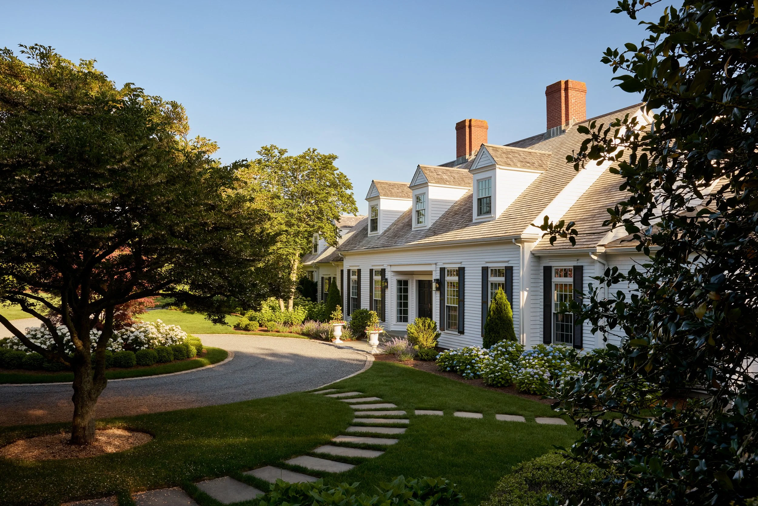 White house with black shutters, surrounded by well-maintained garden and trees, with a curved stone pathway leading up to it, under a clear blue sky.
