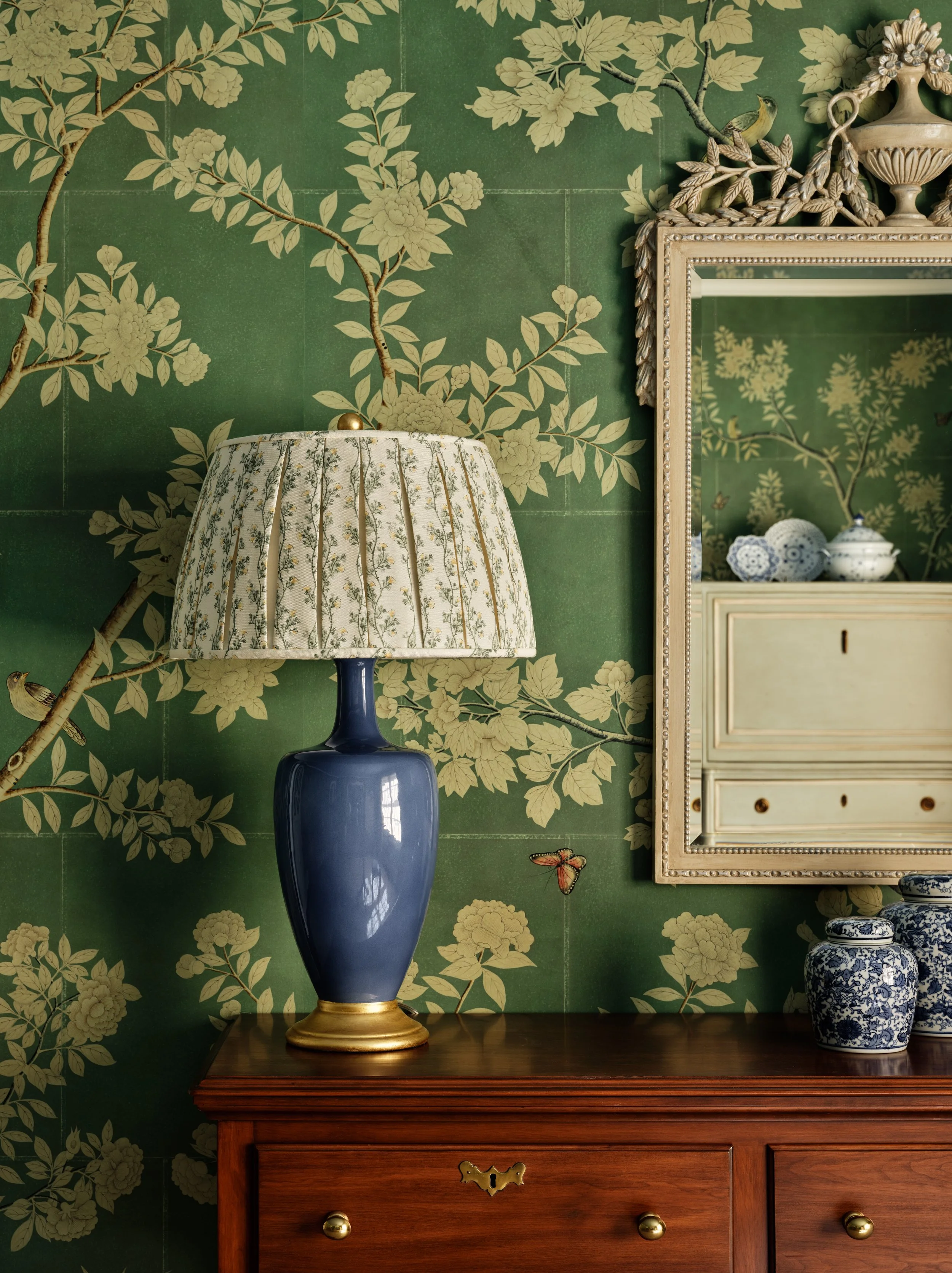 A navy blue ceramic table lamp with a floral fabric lampshade on a wooden surface. Behind, a green wallpaper with a cream floral pattern and butterflies. To the right, a white picture frame and blue and white porcelain vases.