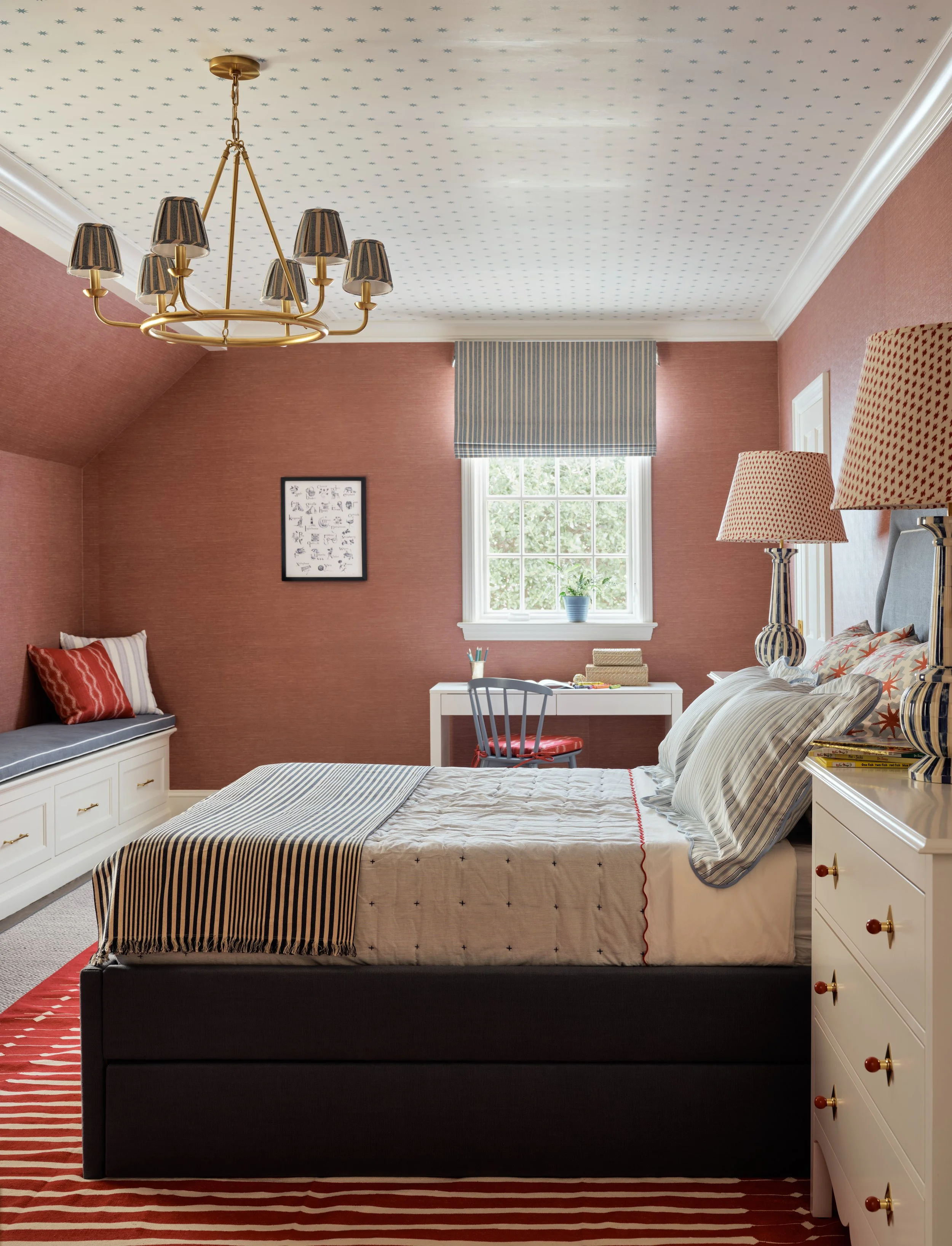 A bedroom with pinkish-red walls, a window with a striped valance, a white desk and chair, a bed with striped and patterned pillows, a white dresser with a striped lamp, a window seat with pillows, and a chandelier hanging from a dotted ceiling.