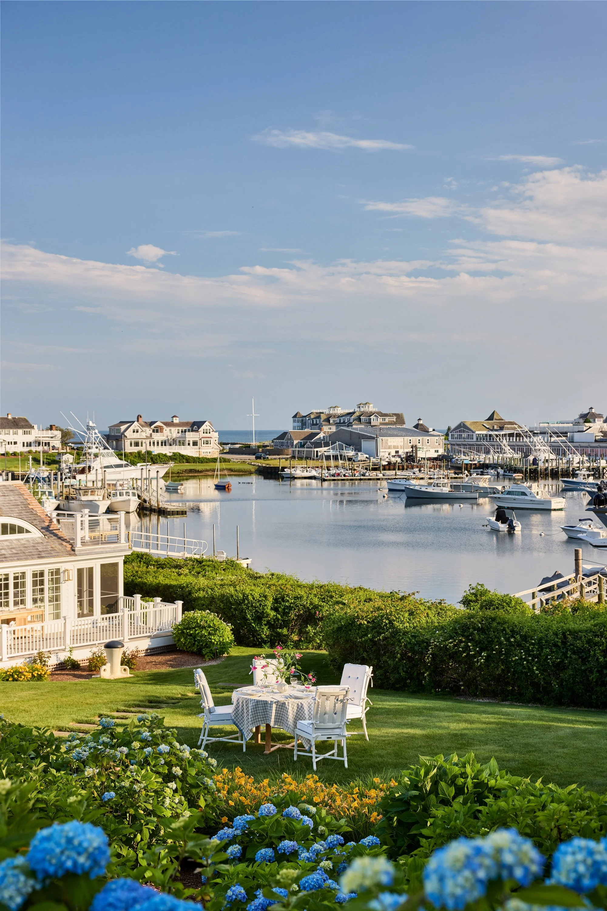A lakeside scene with a small table set for outdoor dining among green bushes and colorful flowers, overlooking a marina with boats and houses under a partly cloudy sky.