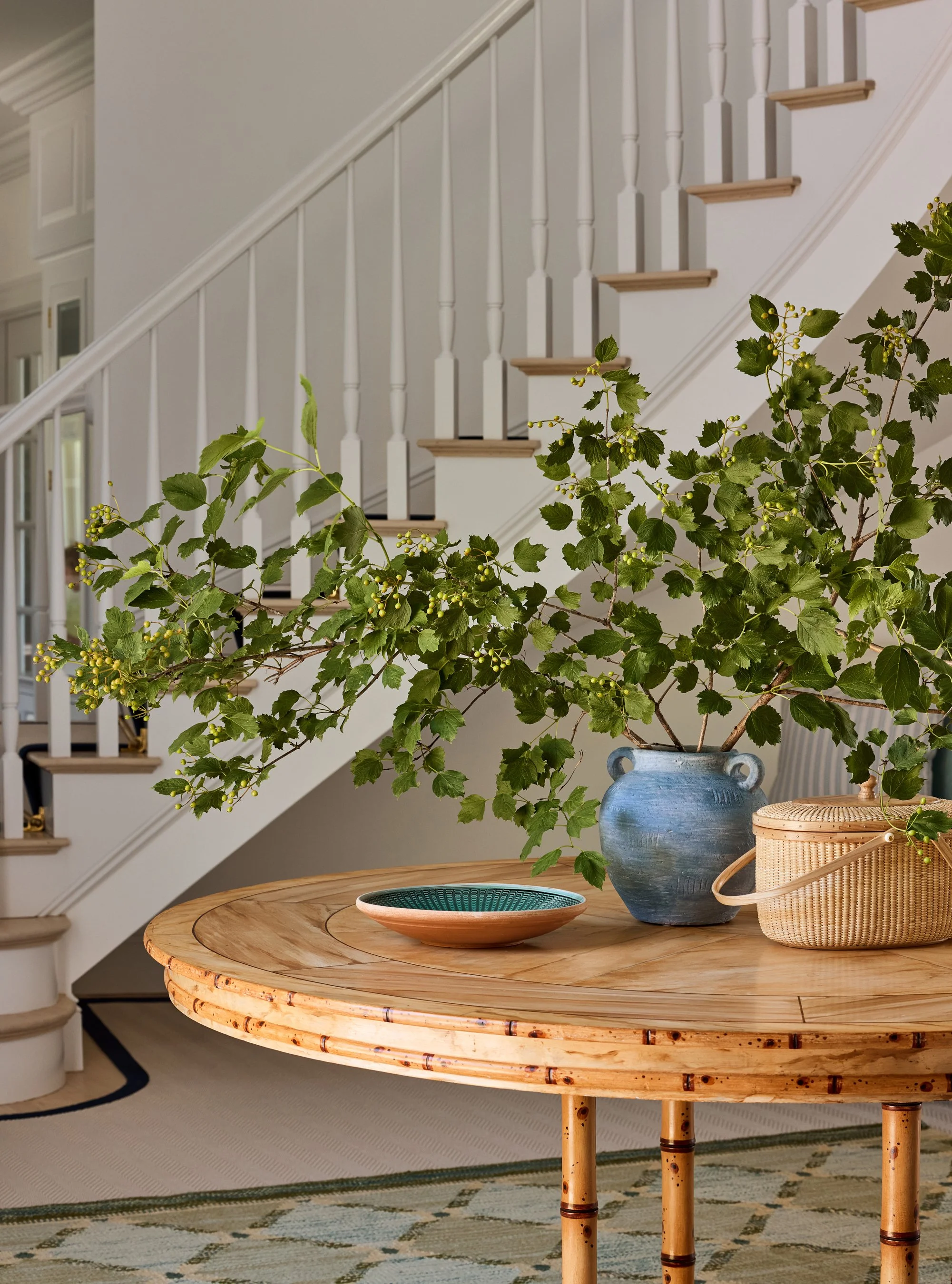 A wooden table with a large green leafy plant in a blue vase, a woven basket, and a teal bowl, with a staircase in the background.