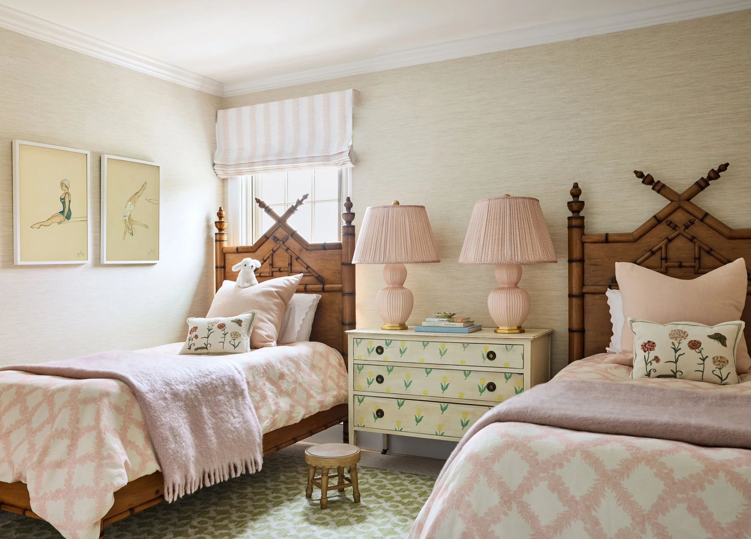 A cozy bedroom with two single beds with wooden headboards, pink bedding, and decorative pillows, a white nightstand with pastel pink lamps, a window with a striped valance, and artwork on the wall.