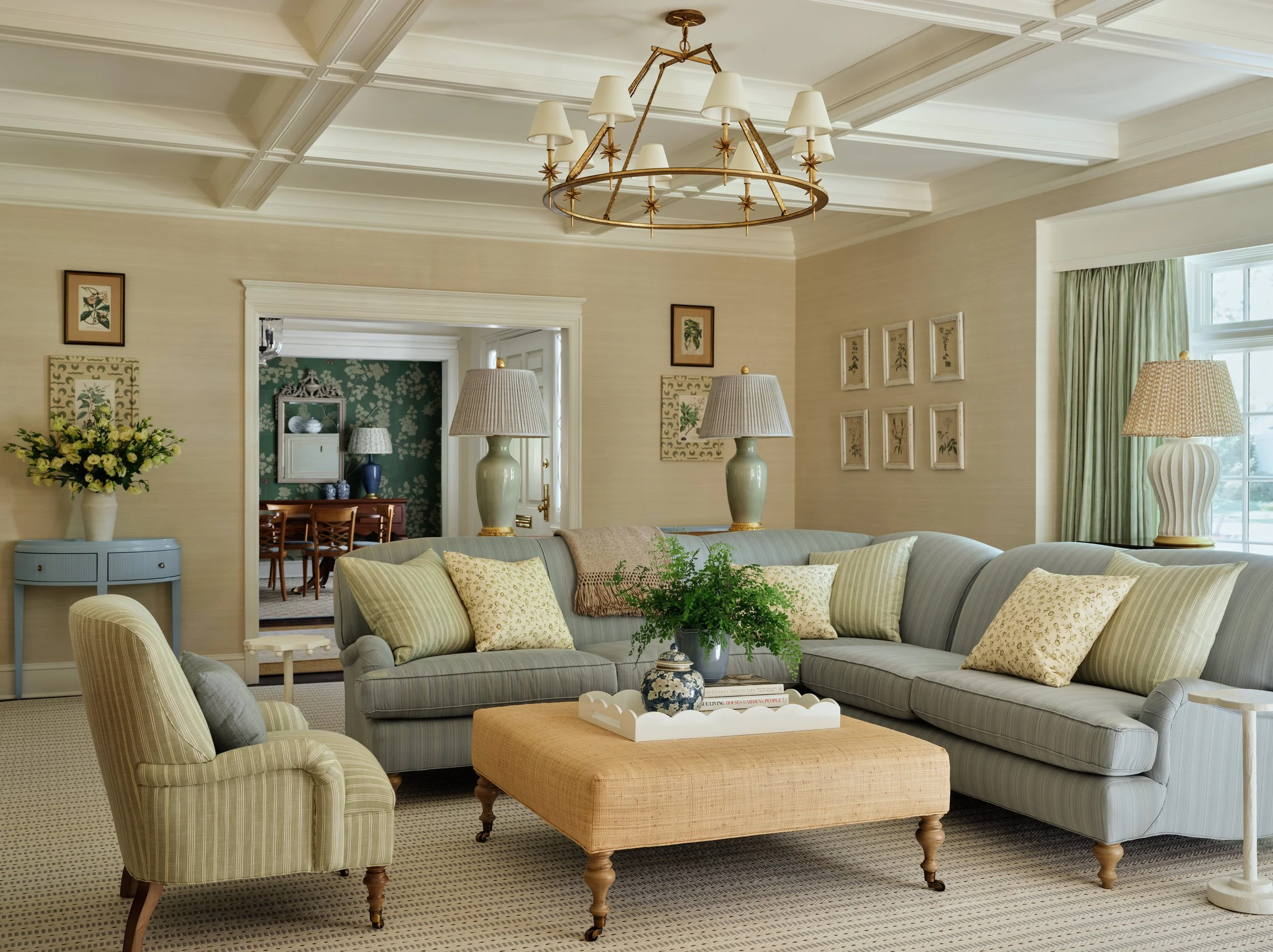 Living room with light-colored sofa, armchair, and an ottoman table with a plant, lamps, and framed artwork, leading to a formal dining room.
