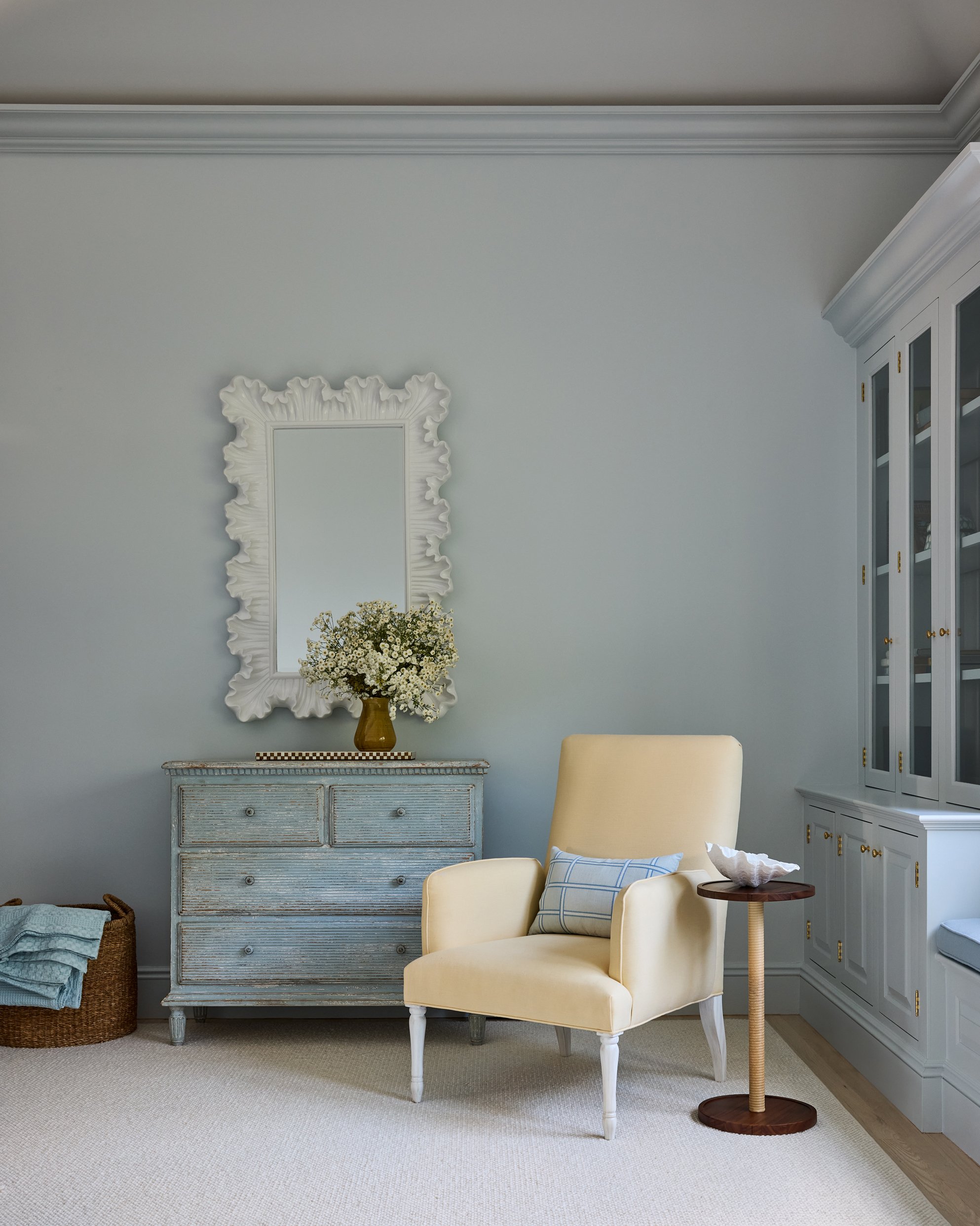 Interior of a cozy living room with a yellow armchair, a small side table, a blue distressed dresser with a vase of white flowers, a mirror with an ornate white frame, a built-in cabinet, and a basket of blankets.