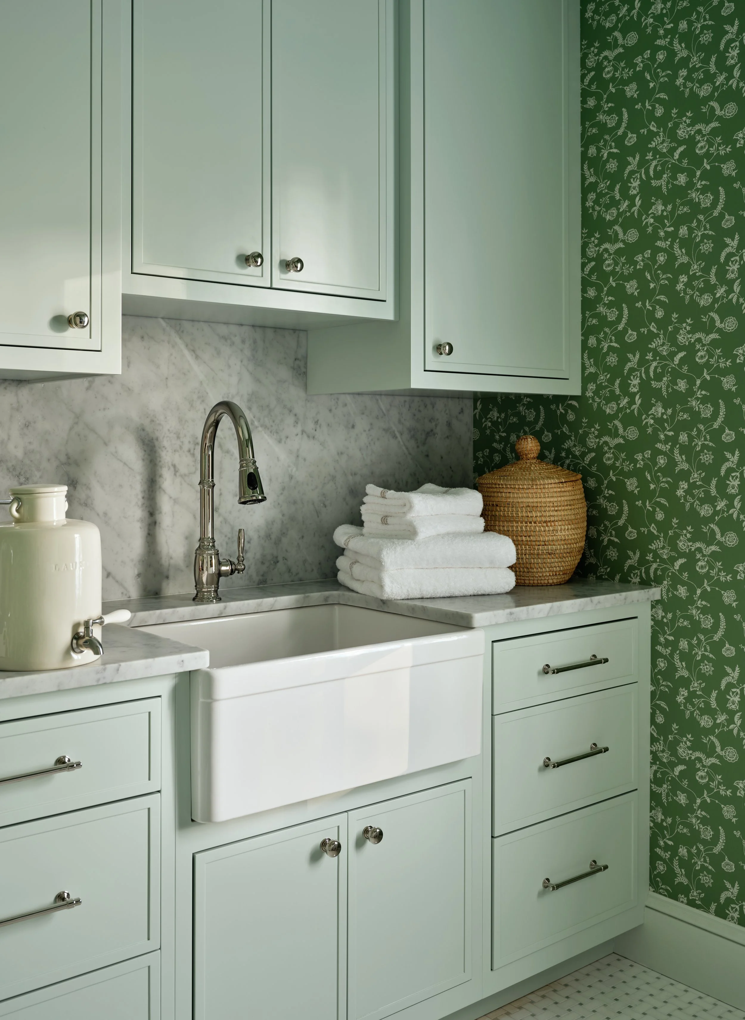 Mint green kitchen cabinets with stainless steel knobs, a white farmhouse sink, marble countertop and backsplash, folded white towels, a wicker jar, and green floral wallpaper.