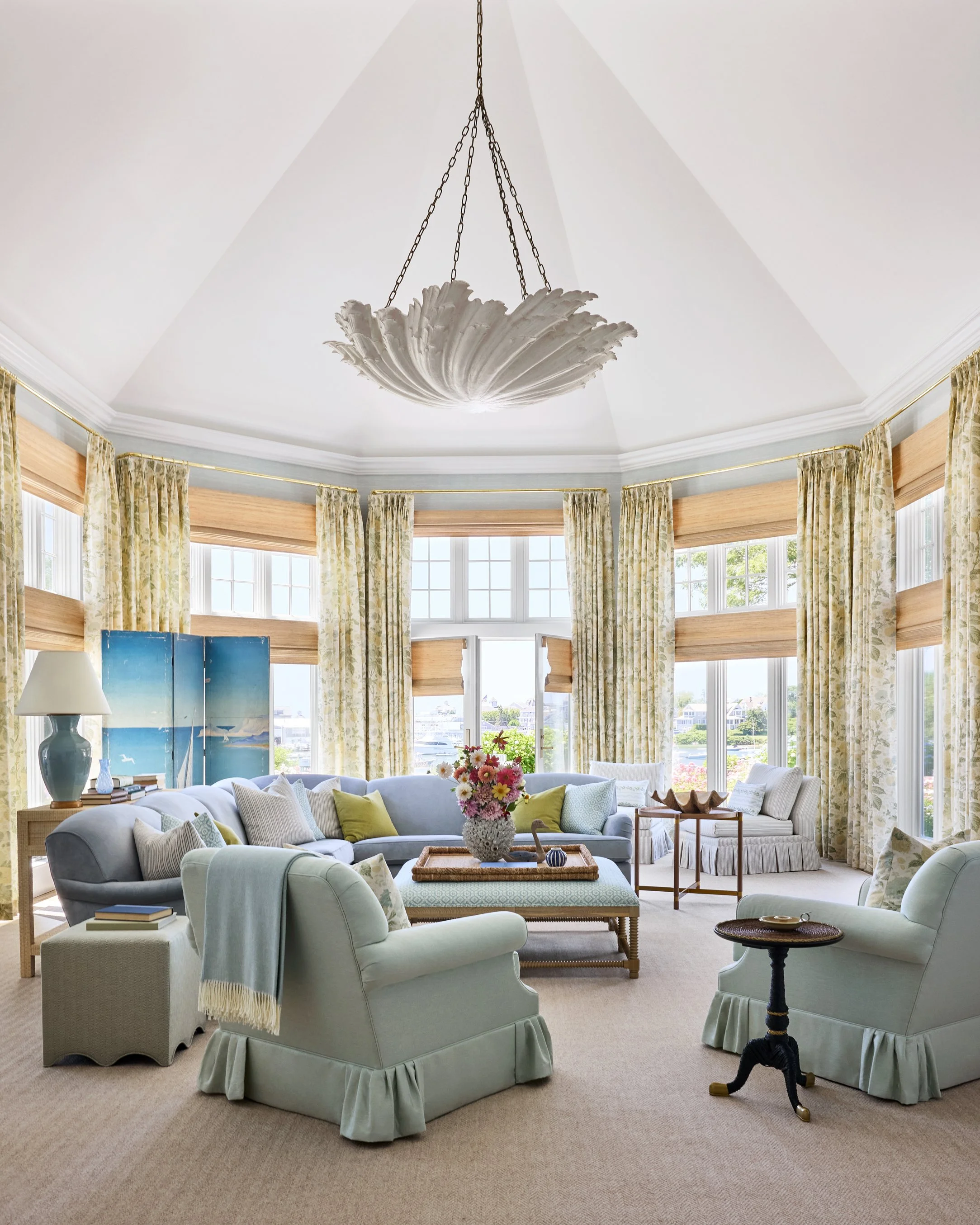 Bright living room with large bay windows, light-colored furniture, floral curtains, and a central coffee table with a vase of pink and white flowers.