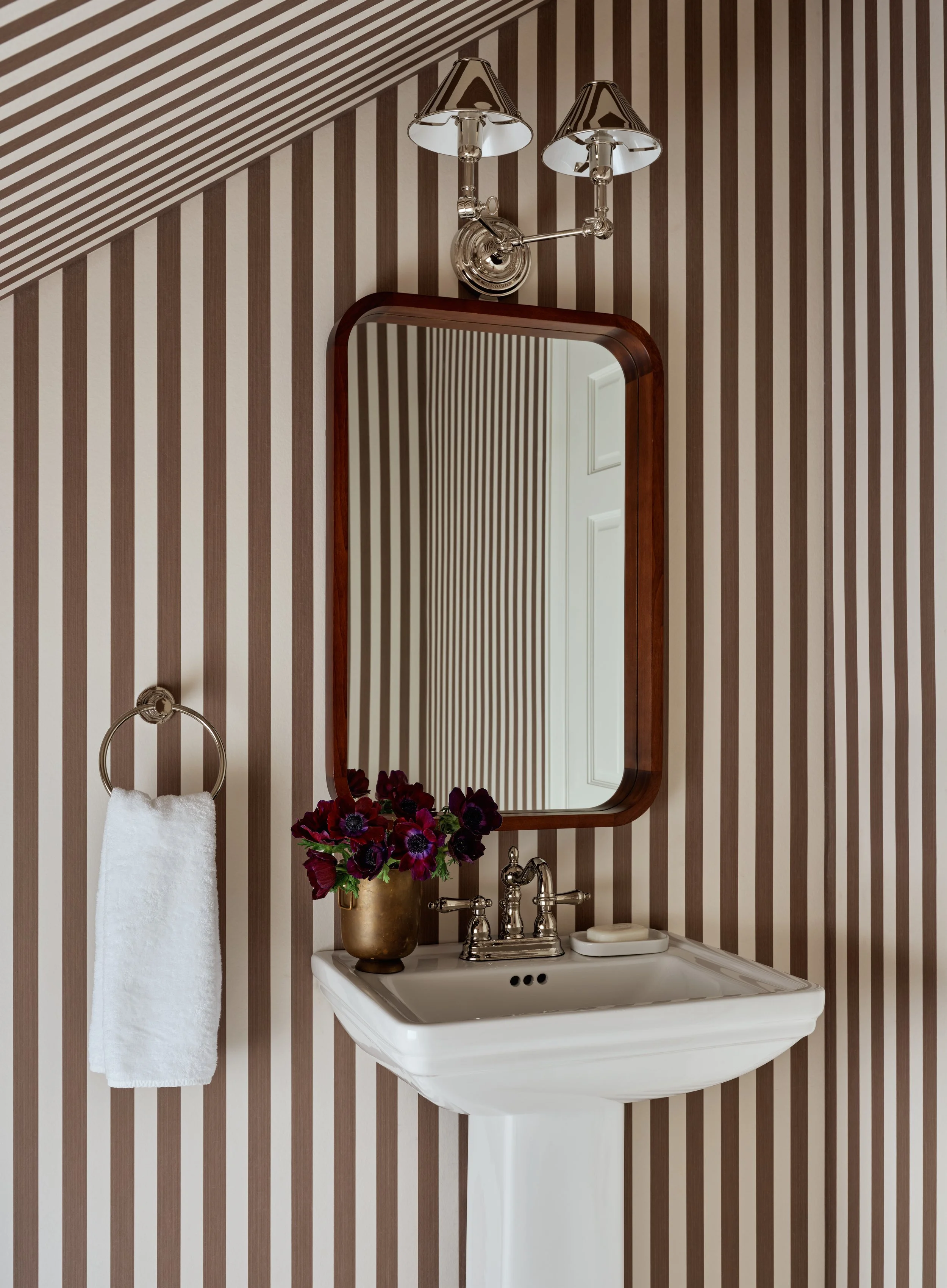 A vintage white pedestal sink with a gold faucet, a mirror with a wooden frame, a brass vase with dark red and purple flowers, a wall-mounted light fixture with two shades, and a white towel hanging on a ring against striped brown and beige wallpaper