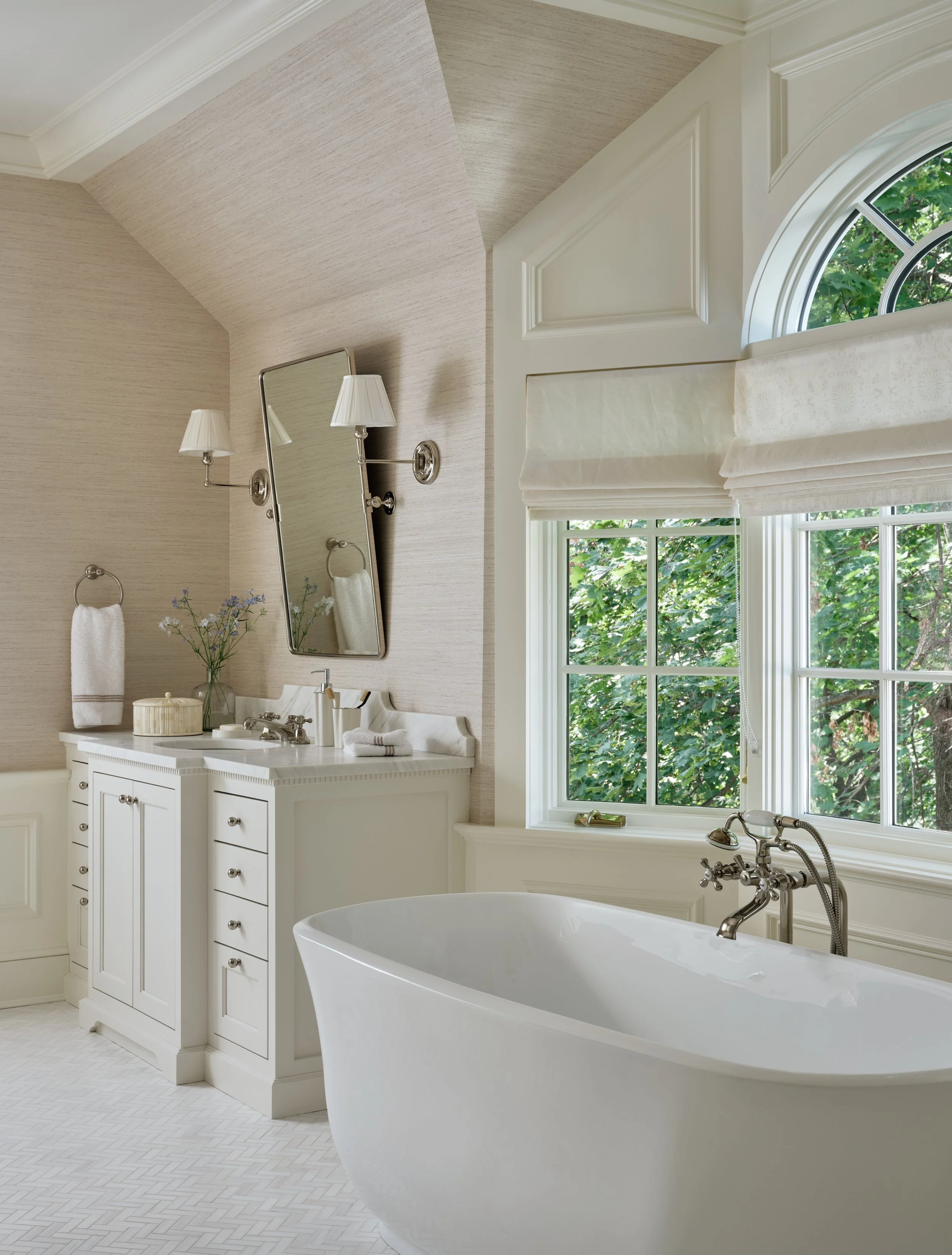 Bright, elegant bathroom with a white clawfoot bathtub, large window with white blinds, and a white vanity with a mirror and wall lamps.