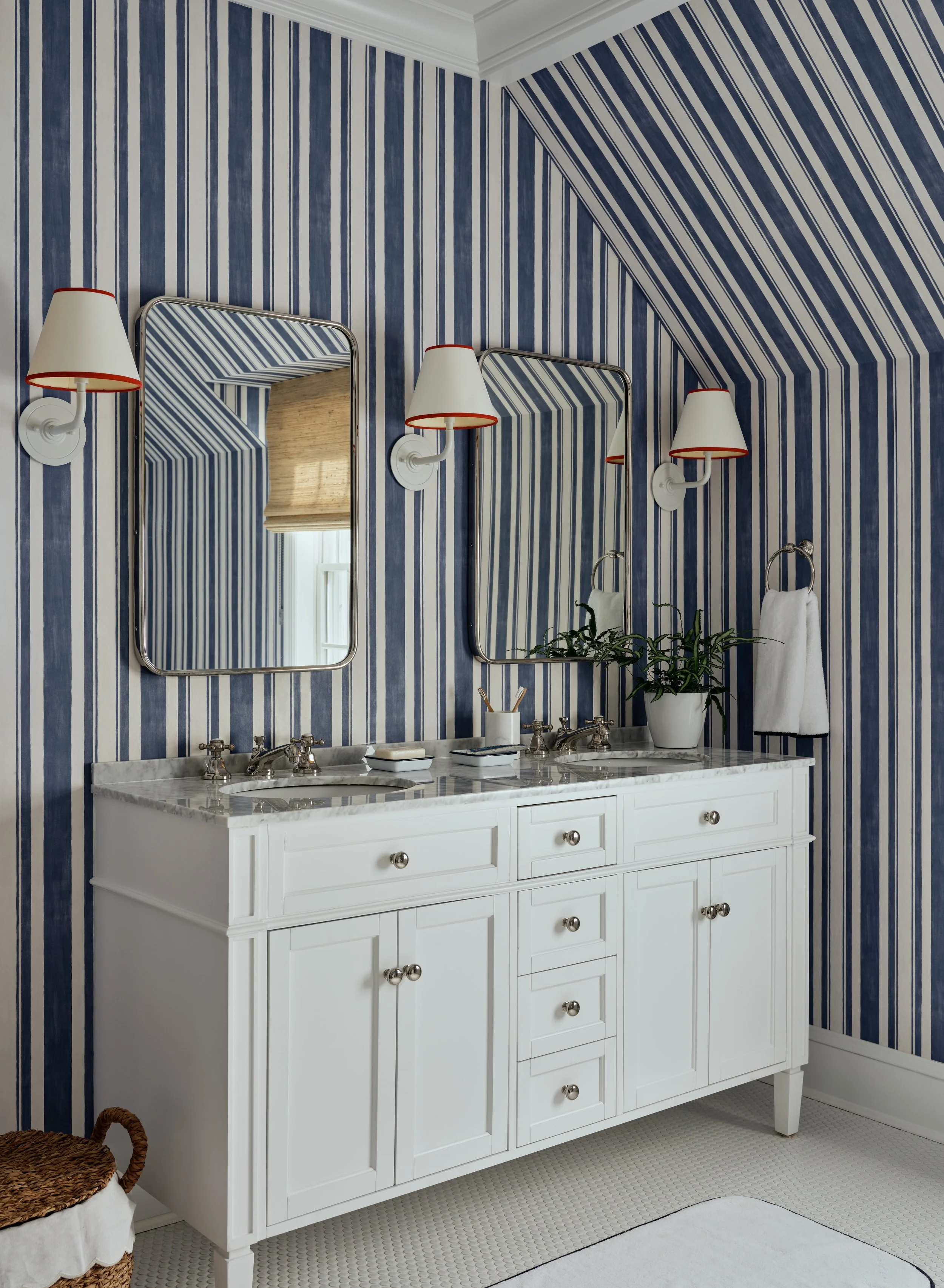 A bathroom with striped blue and white wallpaper, a white double vanity with a marble top, two mirrors, wall-mounted lights, a potted plant, and a towel hanging on a ring.
