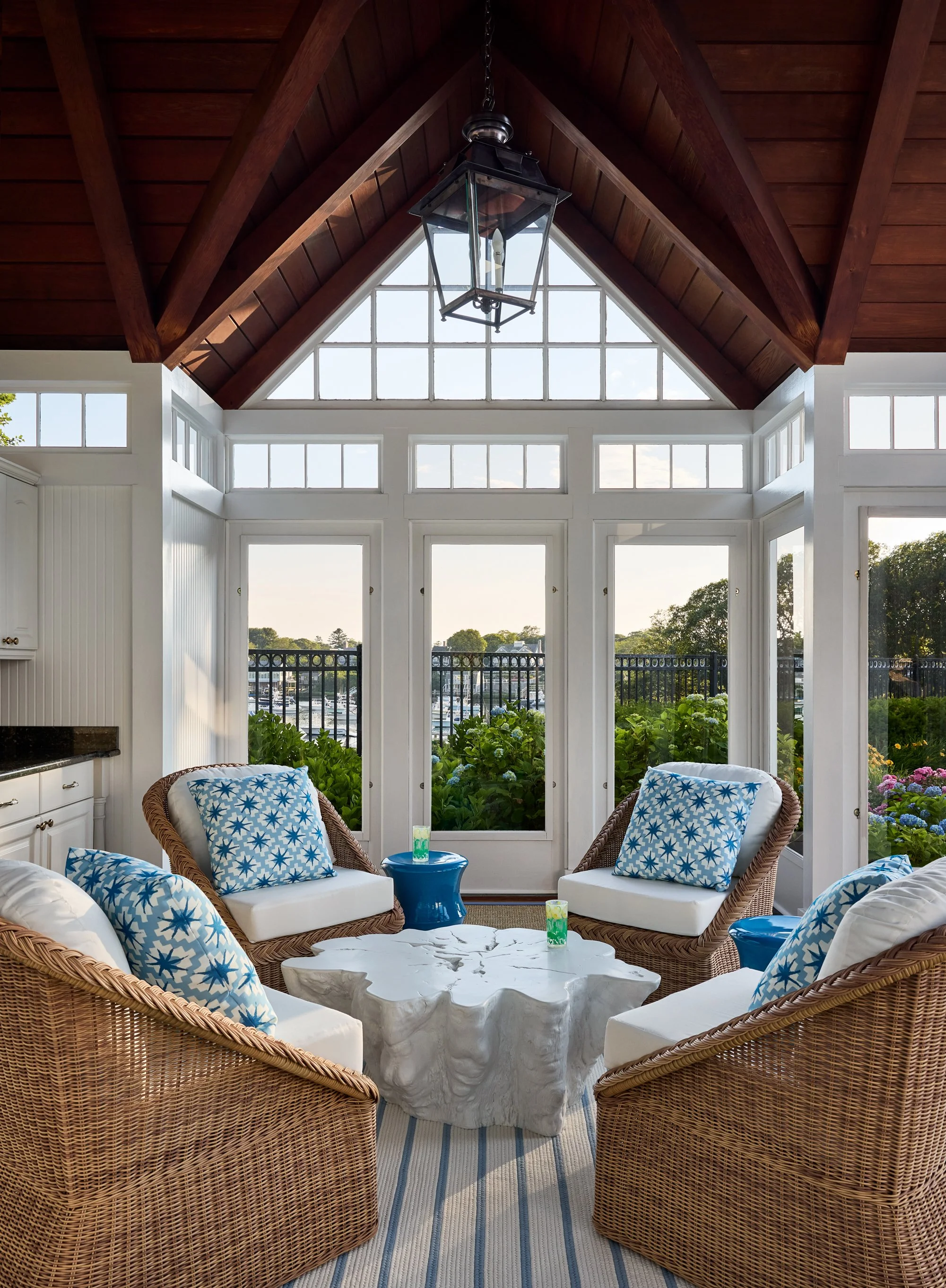 View of a bright sunroom with large windows and a glass ceiling, furnished with wicker chairs with patterned cushions, a white carved coffee table, and small blue side tables, overlooking a garden with flowering plants and a marina in the background.