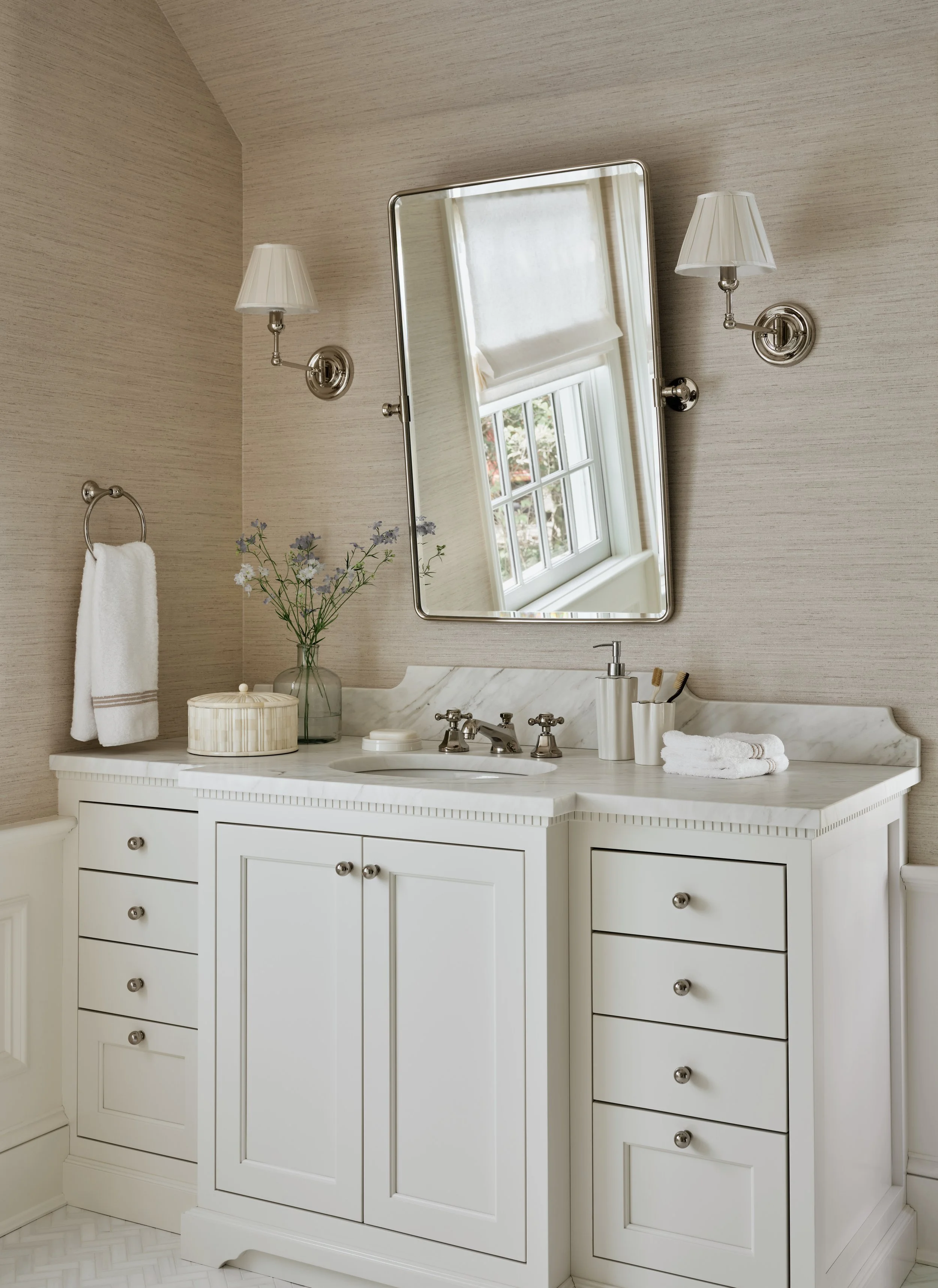 A bathroom vanity with a white cabinet, marble countertop, and a large mirror. There are two wall-mounted lamps, a towel on a ring, a vase with flowers, and bathroom essentials on the counter. A window is reflected in the mirror.