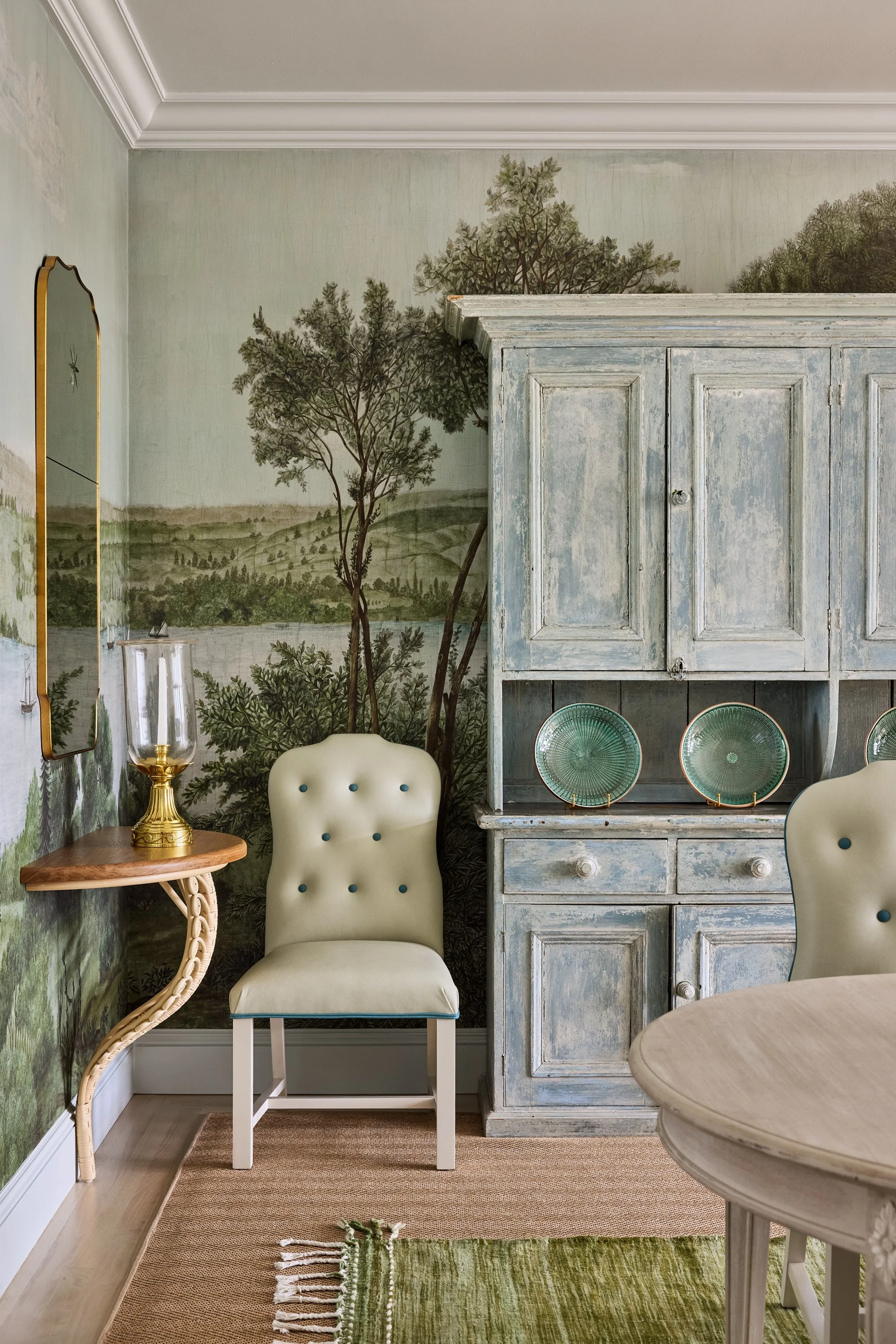 A corner of a room featuring a painted mural of trees and landscape, with a light green chair, a wooden side table with a glass hurricane lamp, a distressed blue cabinet decorated with green plates, a beige round table, and a small green rug.