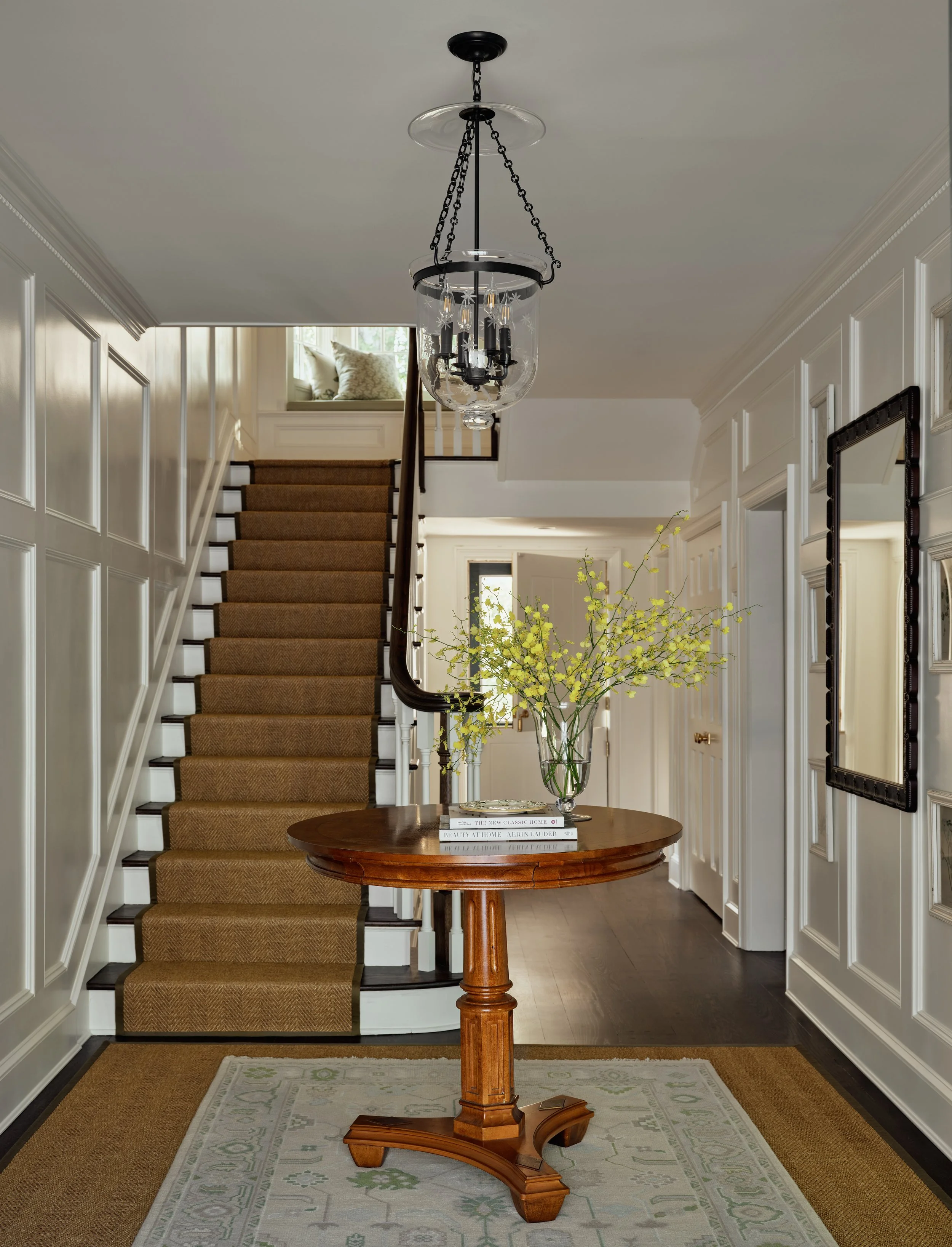 Elegant entryway with a staircase, a wooden table with books and a glass vase of yellow flowering branches, a hanging chandelier, and framed mirror on white paneled walls.