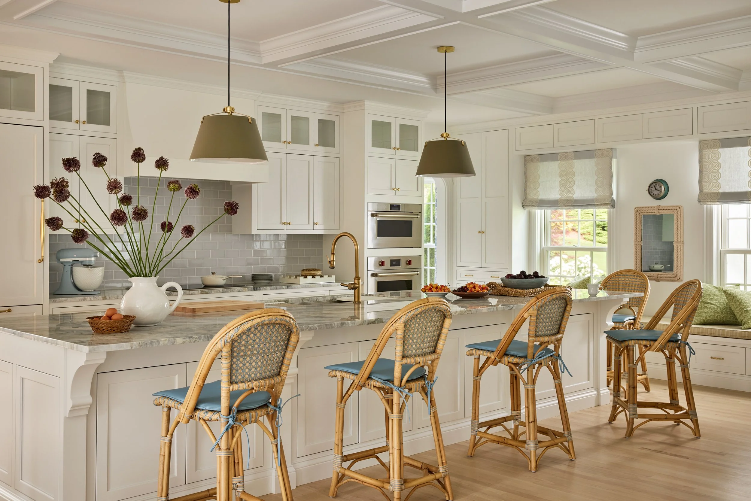 Bright kitchen with white cabinets, marble countertops, gold hardware, and wine-colored accents. Features a kitchen island with a large white vase holding purple allium flowers, four wicker bar stools with blue cushions, and various bowls of fruit. L