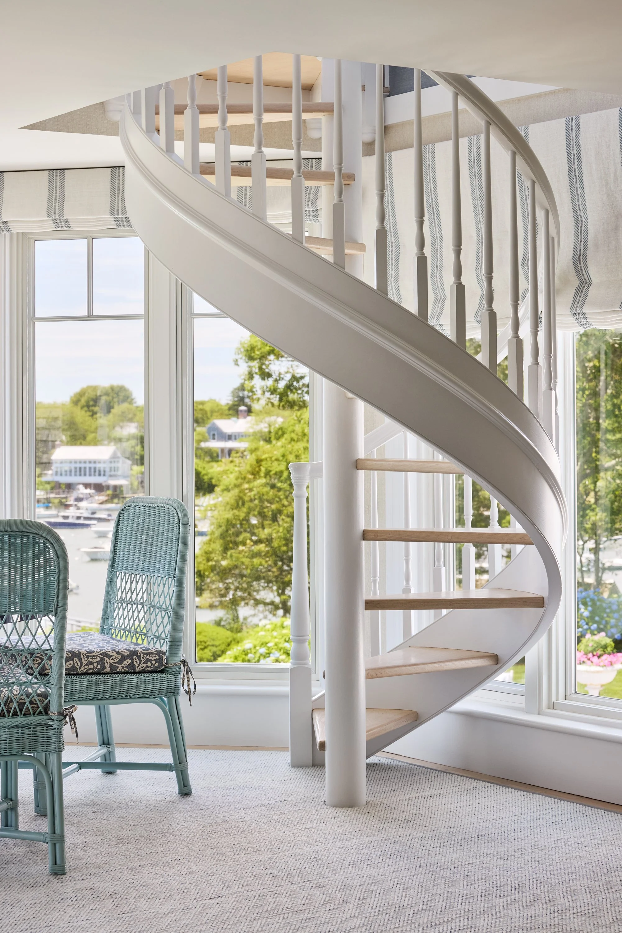 Spiral staircase with white railings and wooden steps inside a bright room with large windows and a view of a harbor with boats and buildings.