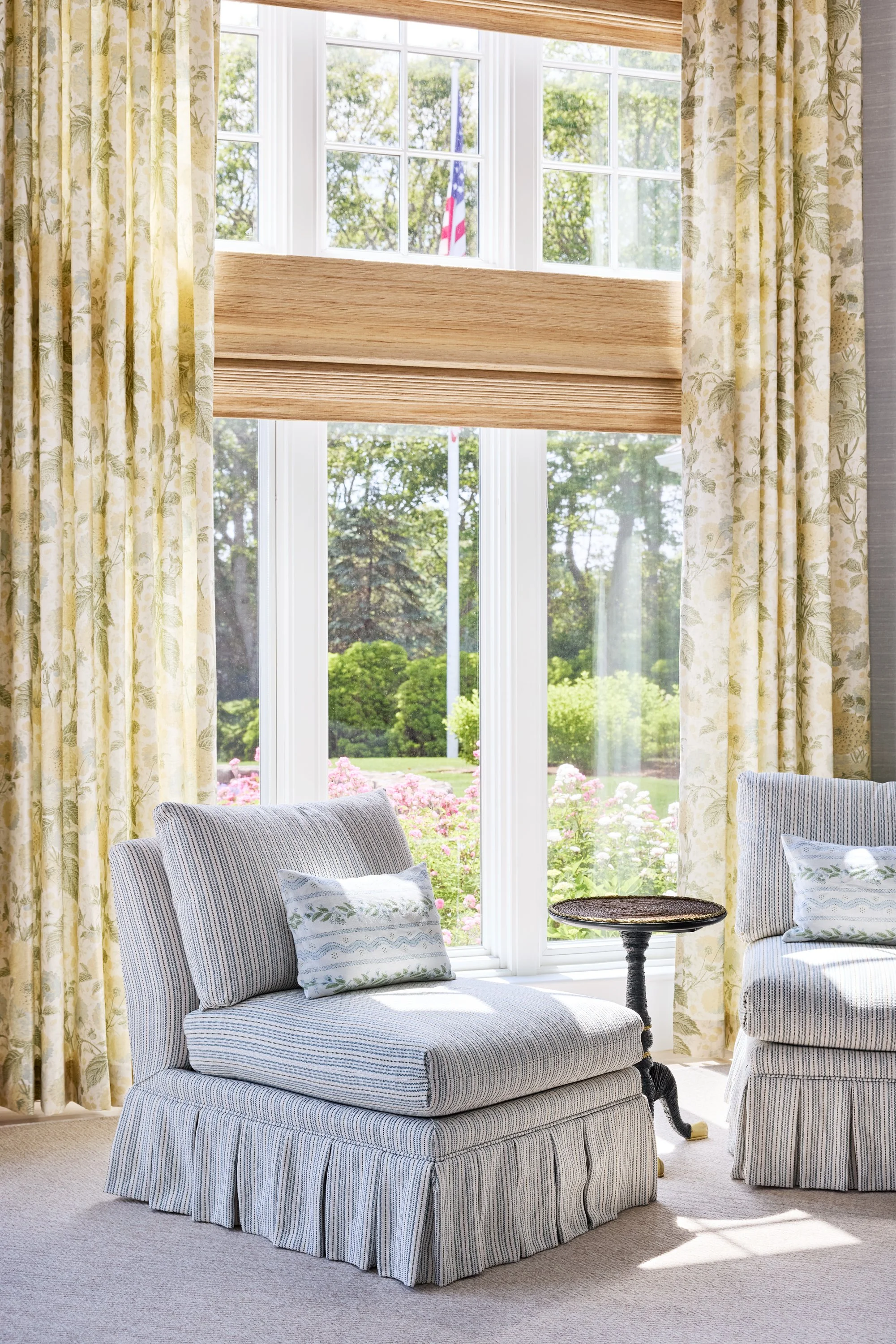 Sunlit living room with striped upholstered chairs, decorative pillows, a small side table, cream carpet, large window with floral curtains, view of greenery and flowers outside.