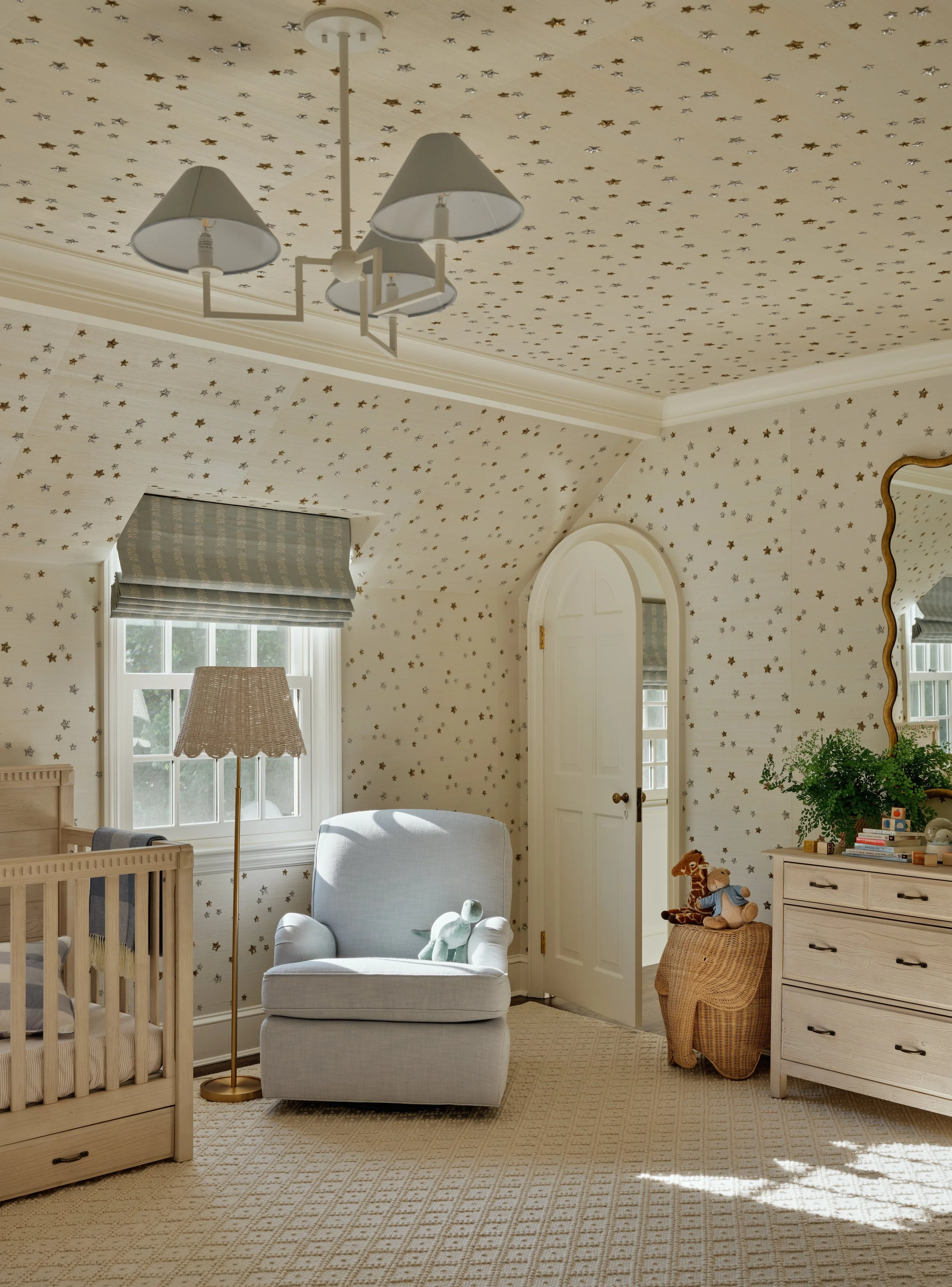 A cozy nursery room with star-patterned wallpaper on the walls and ceiling, a window with a blue-gray Roman shade, a beige crib, a white armchair with stuffed animals, a wicker basket with toys, a dresser with a plant and toys, and a ceiling light fi