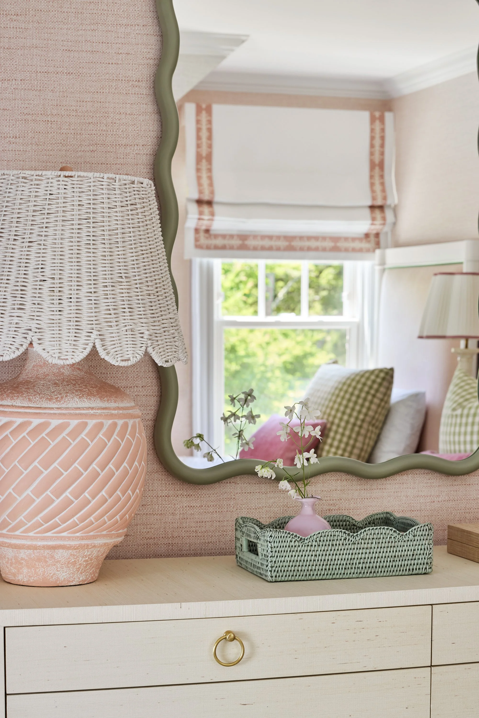 Close-up of a pink decorative lamp, a small pink vase with white flowers, and a mirror reflecting a cozy bedroom with pillows, window, and soft pink walls.