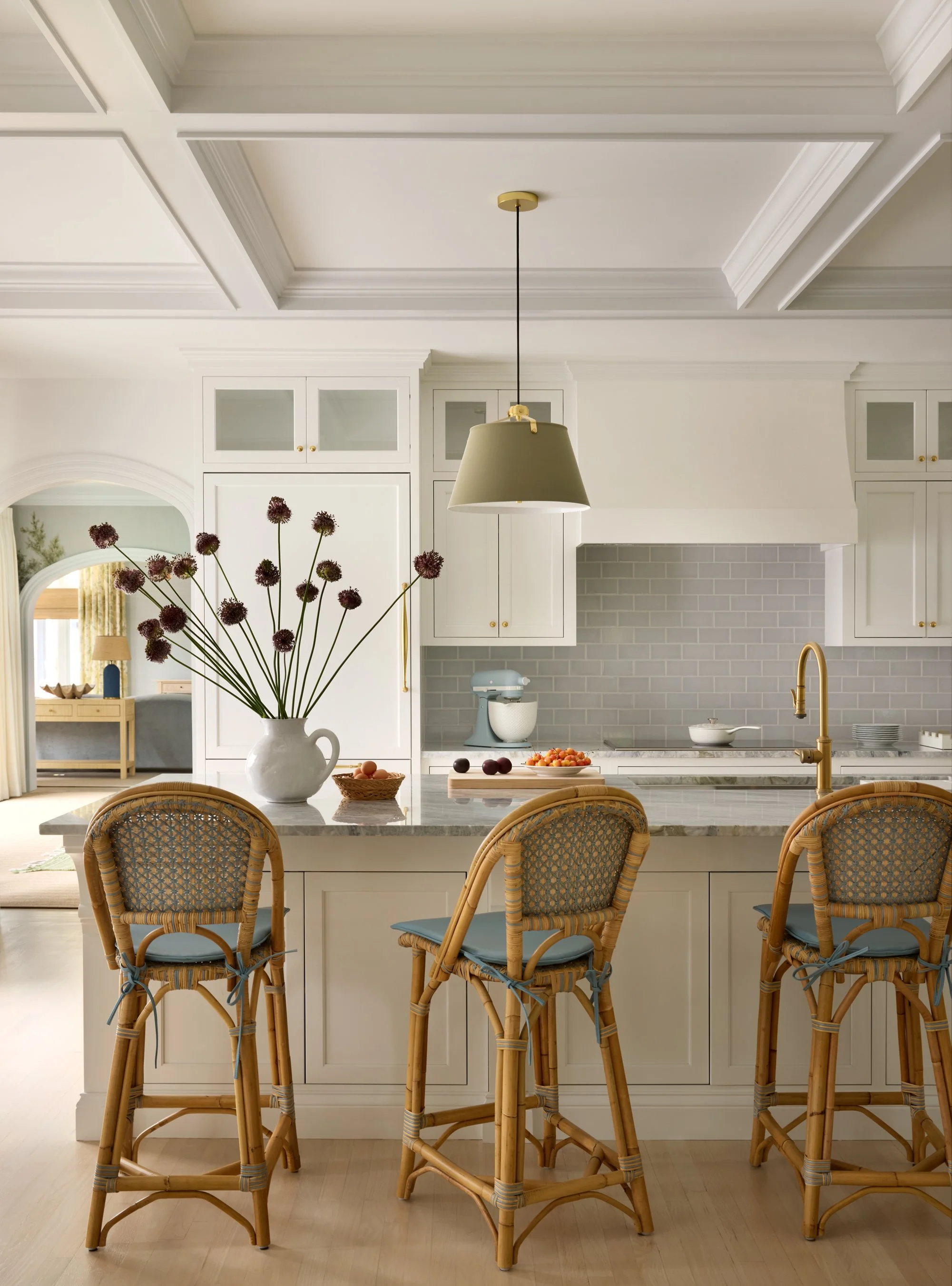 Bright kitchen with white cabinets, gray tile backsplash, marble countertop, brass faucet, hanging light fixture, basket of fruit, large white vase with purple flowers, and three rattan bar stools with blue cushions.