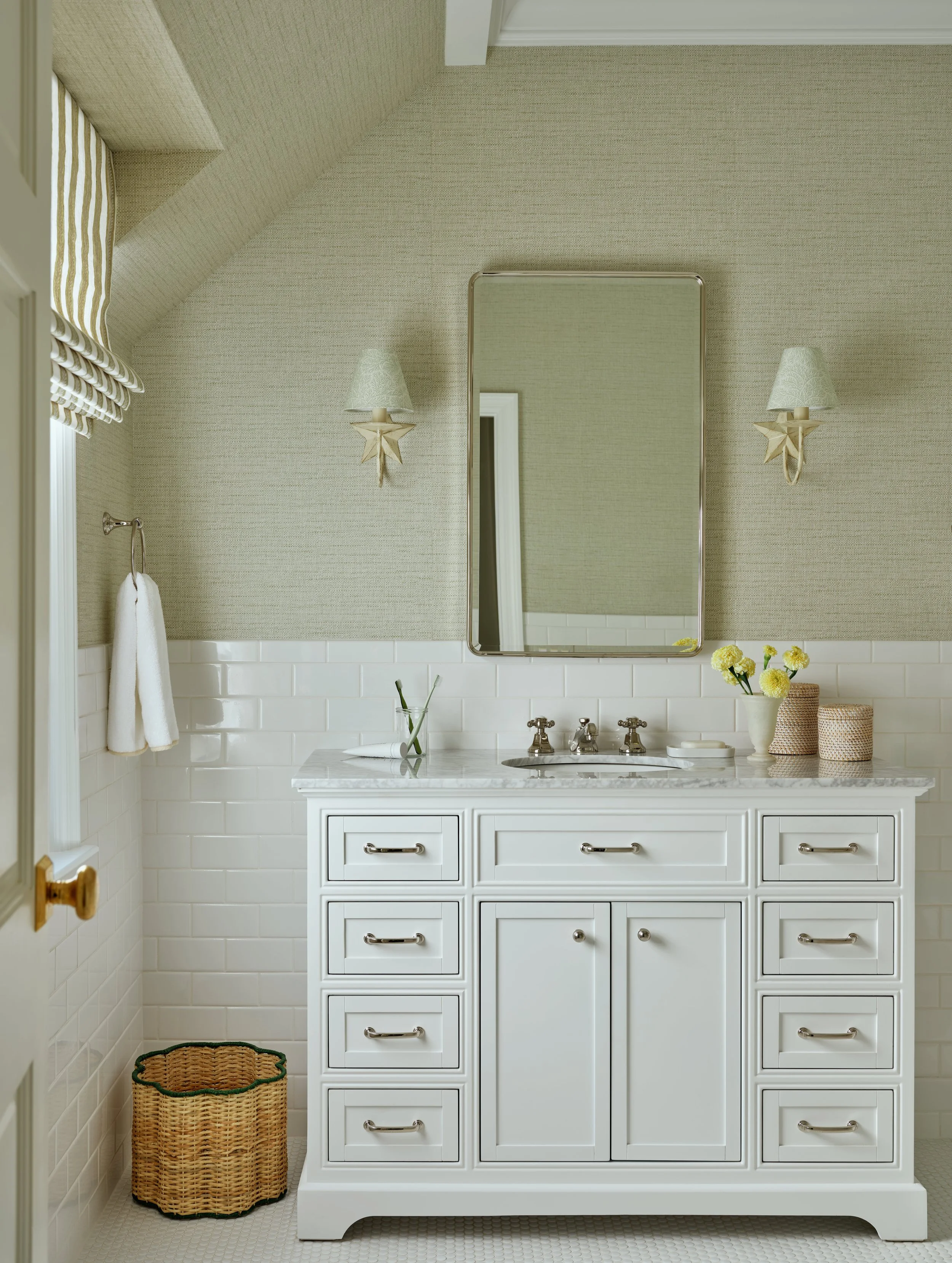 A bathroom vanity with a white cabinet, marble countertop, and a mirror. There are two wall sconces with star-shaped ornaments, a vase with yellow flowers, and two woven baskets on the counter. A towel hangs on a hook on the wall, and a wicker laundr