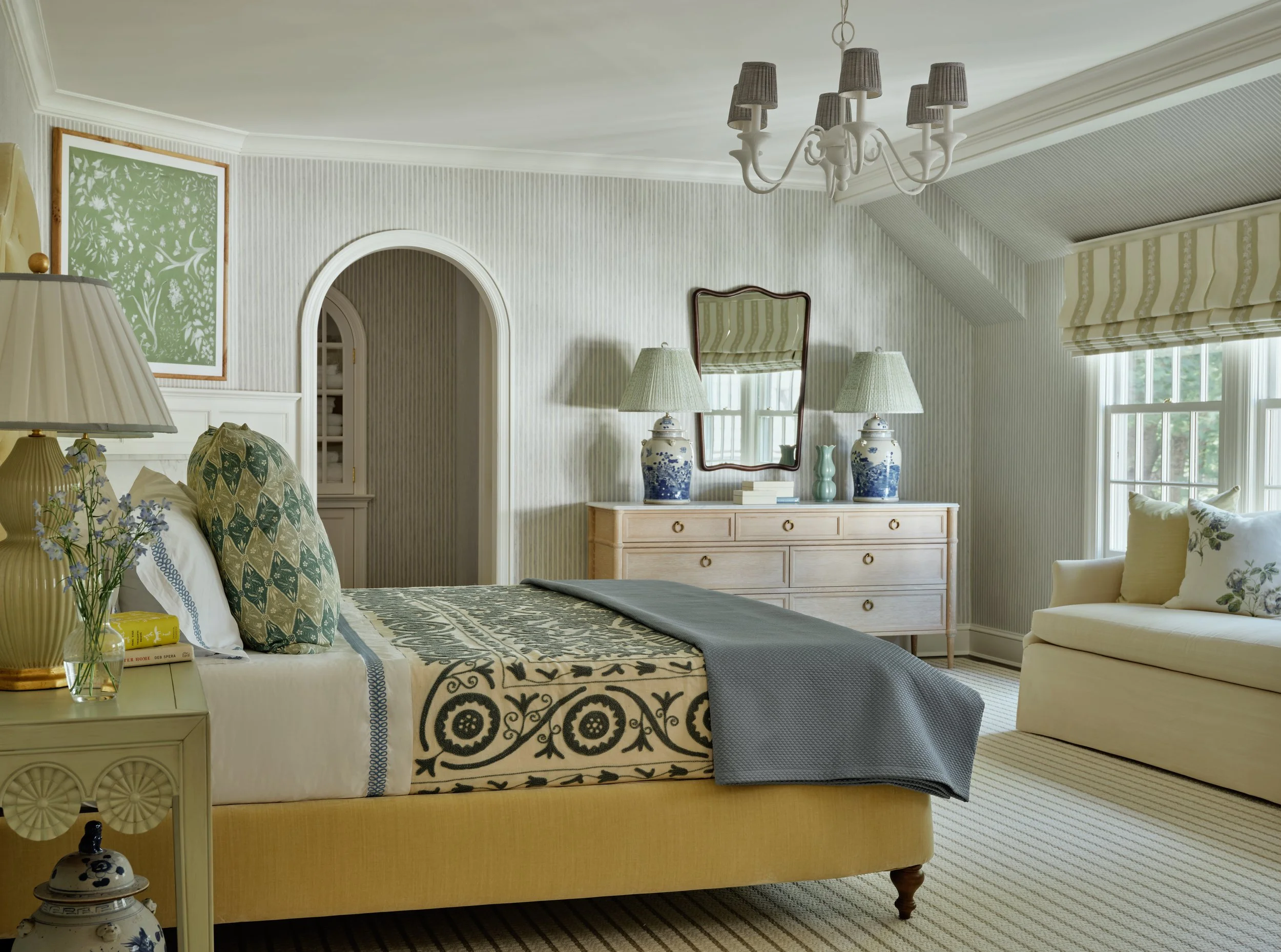 A bedroom with a yellow bed, patterned pillows, a bedside table with a lamp and flowers, a dresser with two lamps and a mirror, a cream sofa with cushions, and a window with striped curtains.