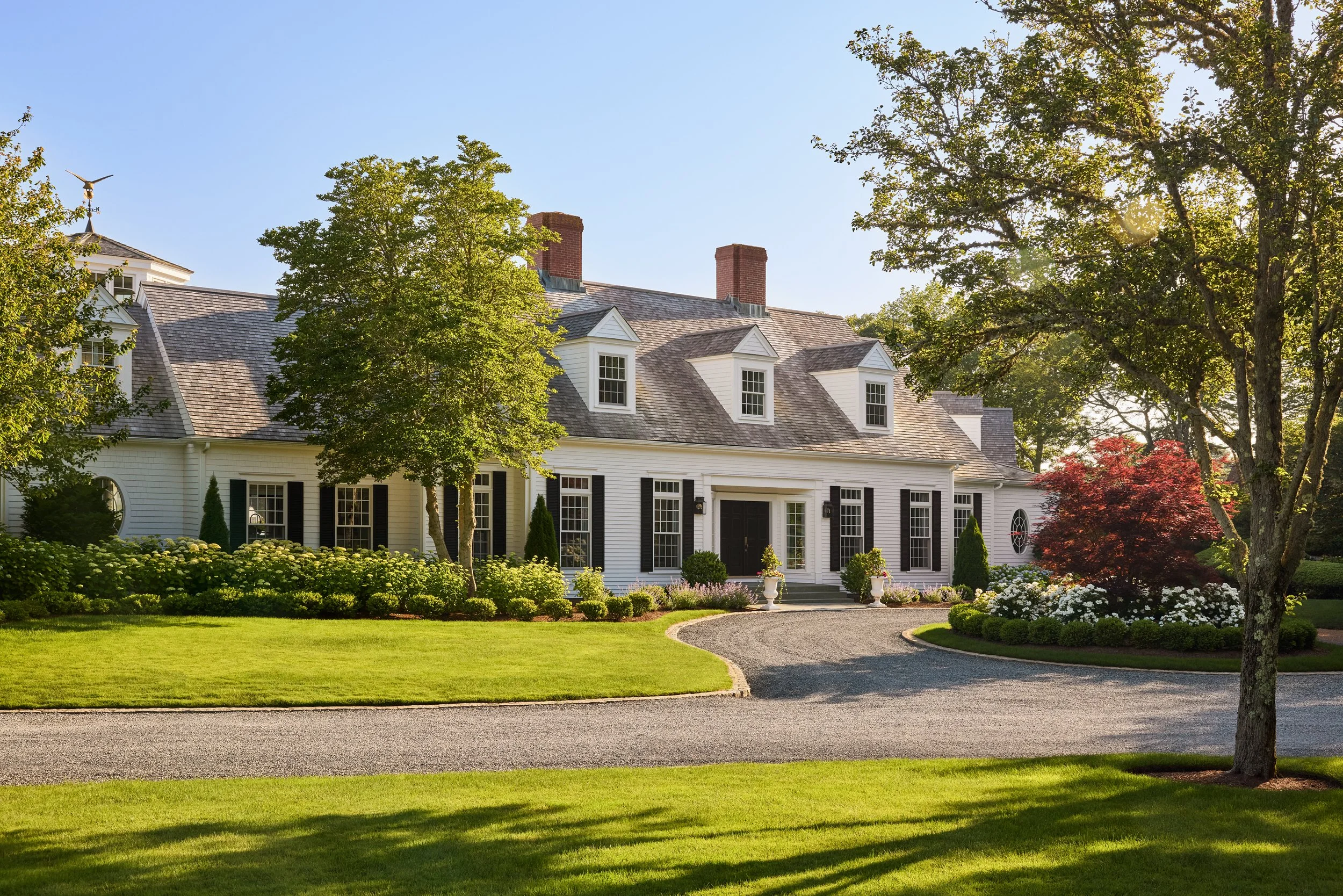 A large white house with black shutters, multiple chimneys, and a curved driveway surrounded by a well-manicured lawn and garden with trees and shrubs.