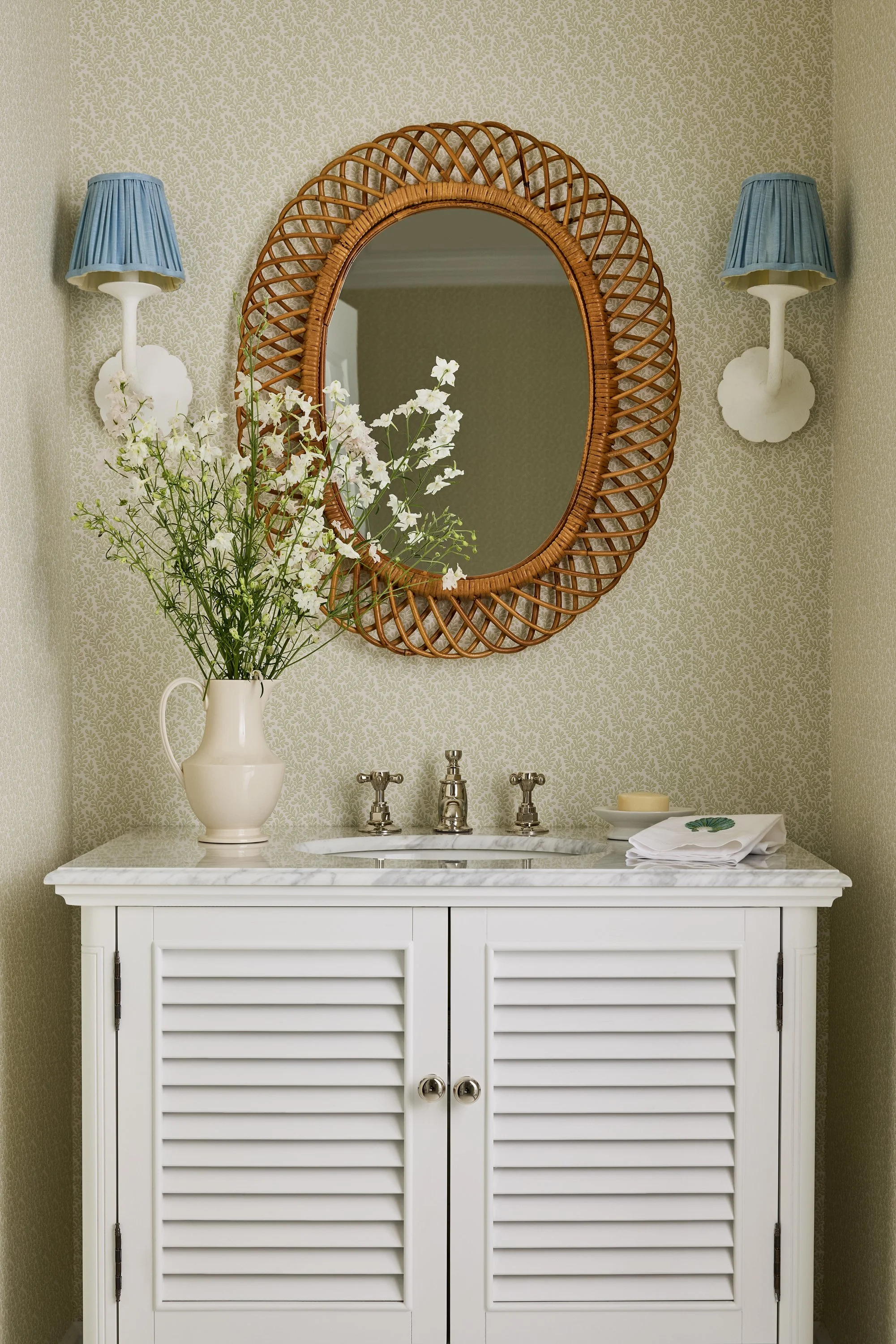 Bathroom with white cabinet, marble countertop, oval mirror with rattan frame, two wall sconces with blue lampshades, white vase with flowers, soap, and towels on the countertop.