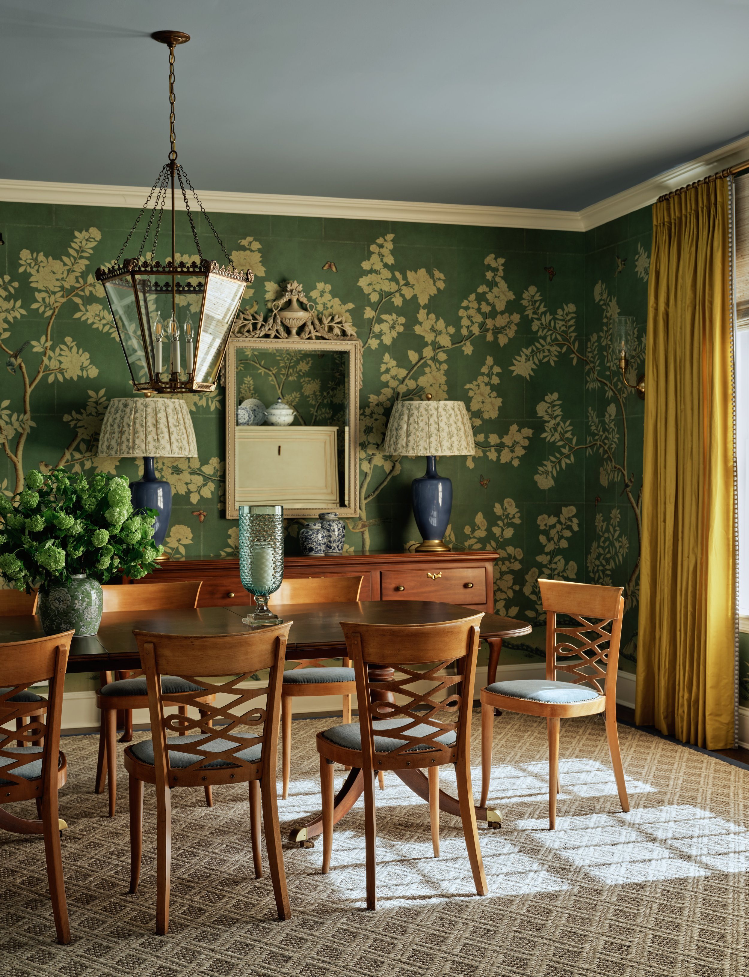 A dining room with a round wooden table and six matching chairs with cushioned seats, a large vase of green flowers, and a decorative glass vase. The room has dark green wallpaper with a floral and butterfly pattern, two blue table lamps with floral 