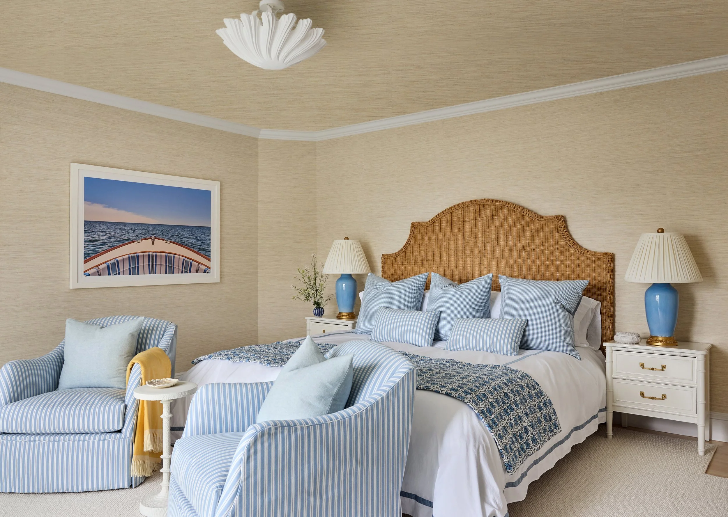A cozy bedroom with a king-sized bed with a rattan headboard, blue and white pillows, and a blue and white quilt. There are two white nightstands with blue lamps, a chair with light blue striped upholstery, and a framed picture of a boat on the wall.