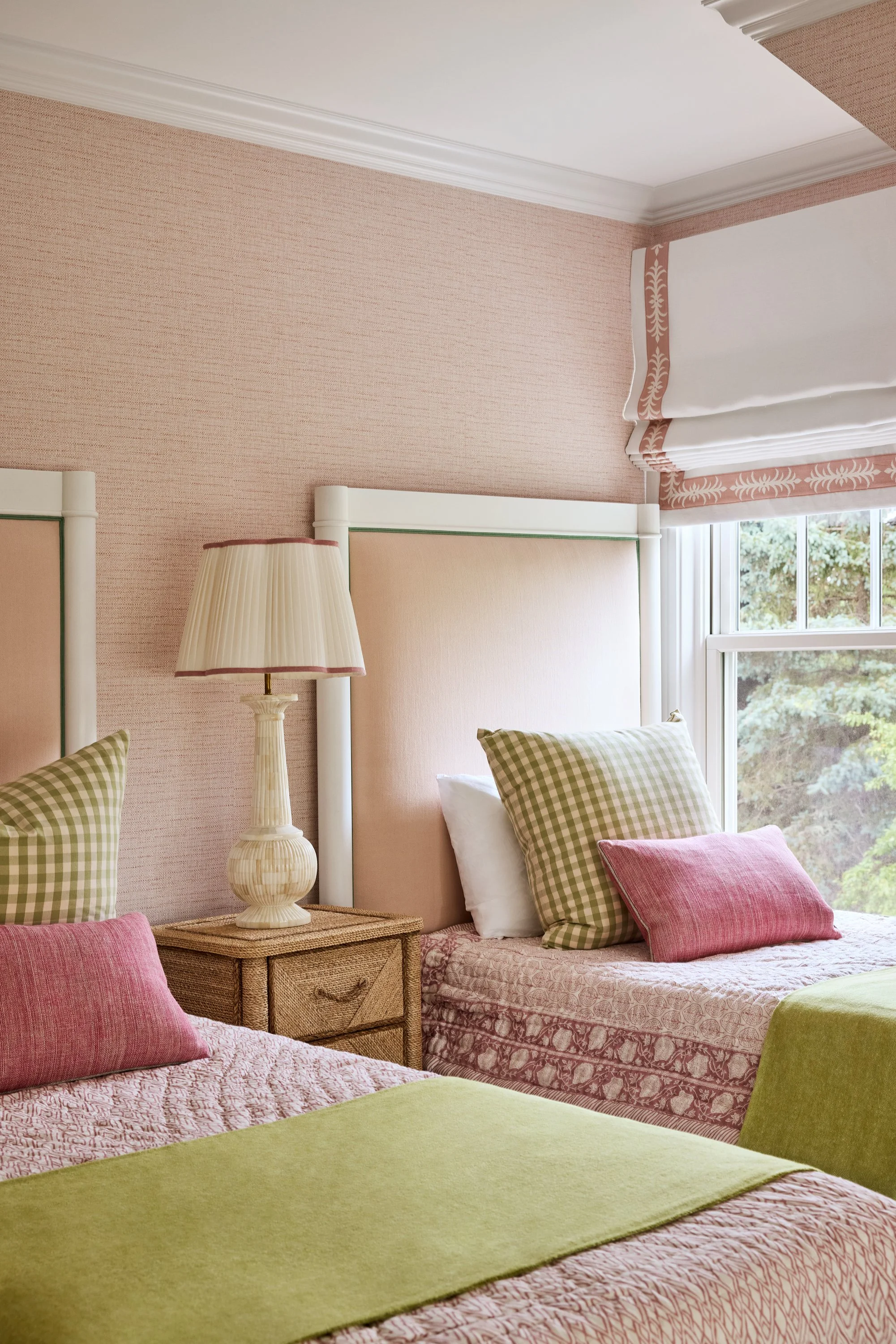 Two beds with pink and green bedding and checkered pillows, a wicker nightstand with a beige lamp, pink textured wallpaper, a window with white and pink curtains, and greenery outside.