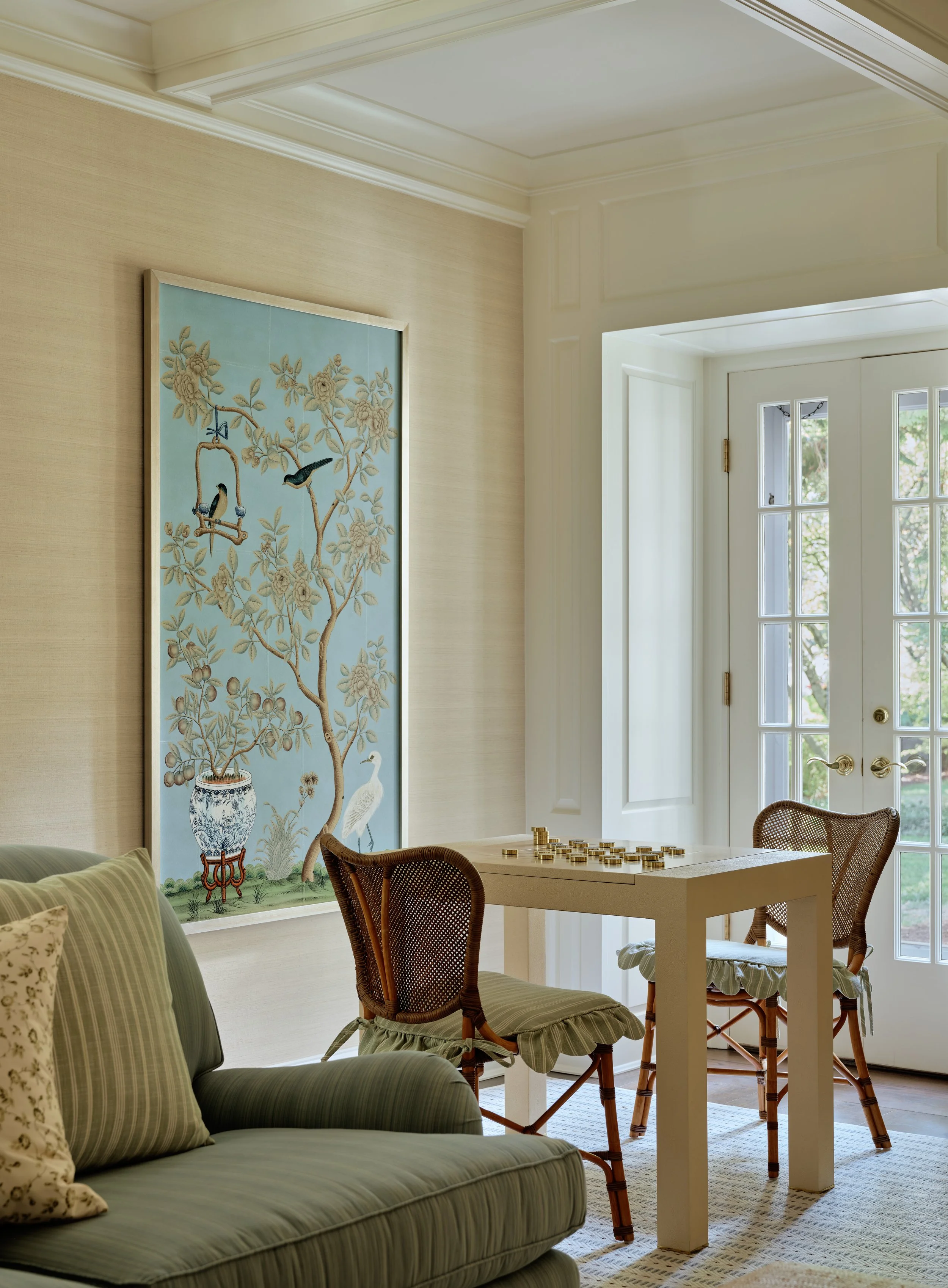 A cozy living room corner with a green striped sofa, a white table with a checkers game, two wicker chairs with green cushions, a large framed artwork featuring birds and a vase, and a door with glass panels leading to a garden.