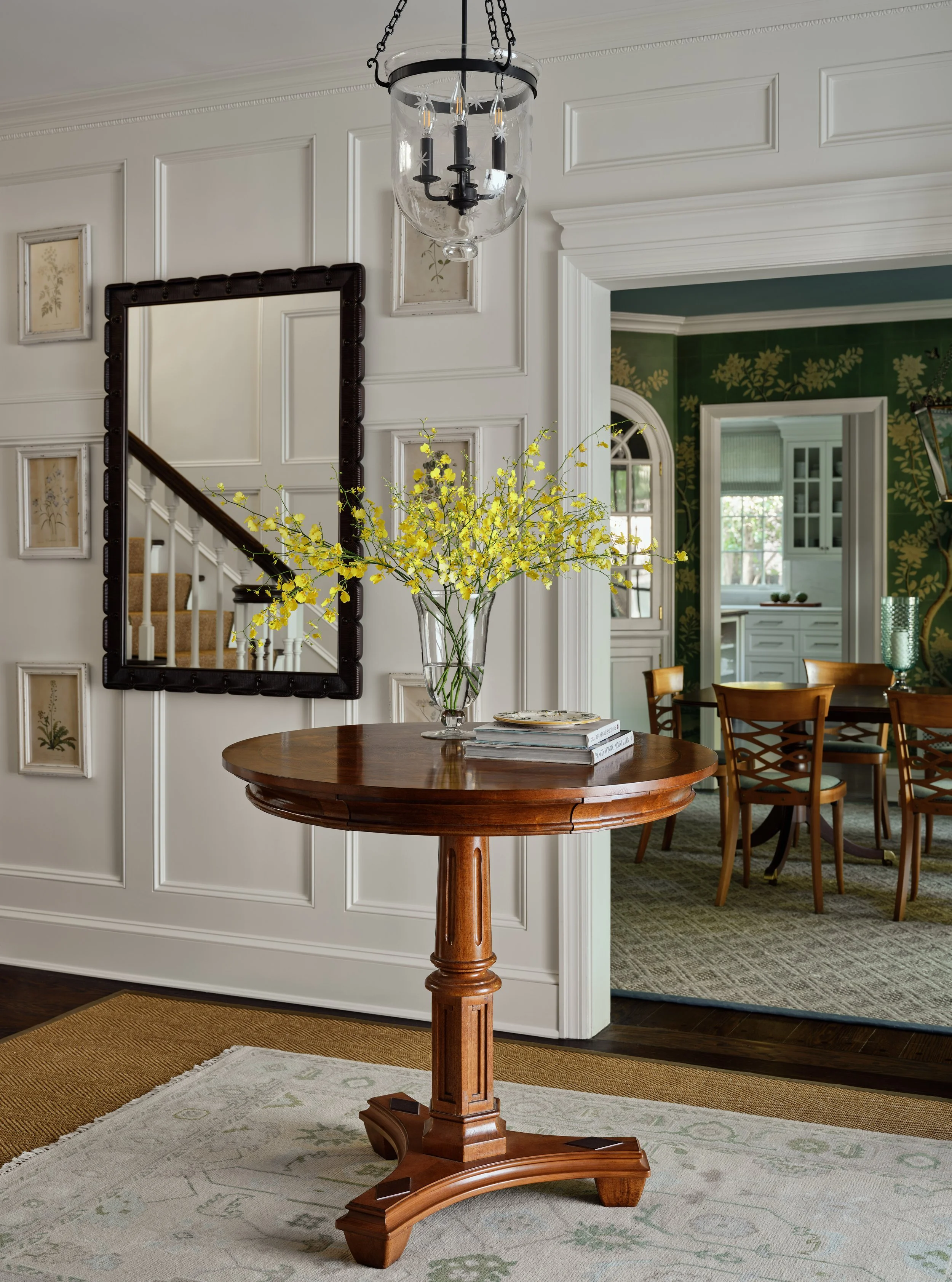 A wooden round table with a vase of yellow flowers and a few books in a home dining area. There is a mirror on the wall, a chandelier above, and an opening to a kitchen or another room with dark green walls and wooden dining chairs.