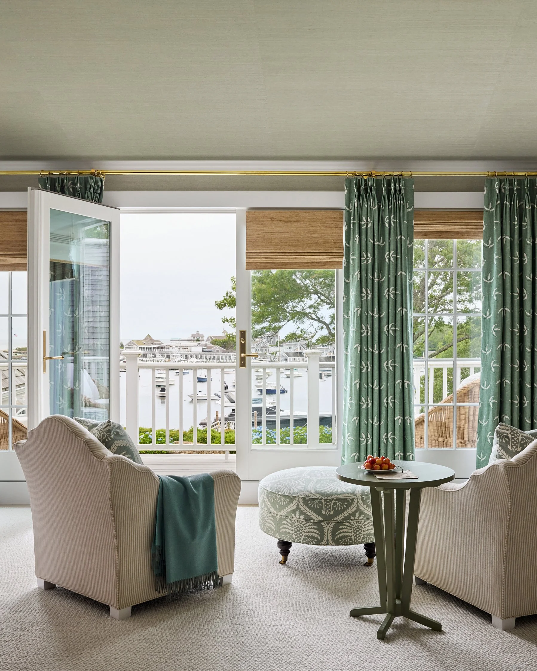 Living room with two beige armchairs, a round ottoman, and a small green side table with a bowl of cherries. Large windows and open door leading to a balcony overlooking a marina with boats and waterfront houses.