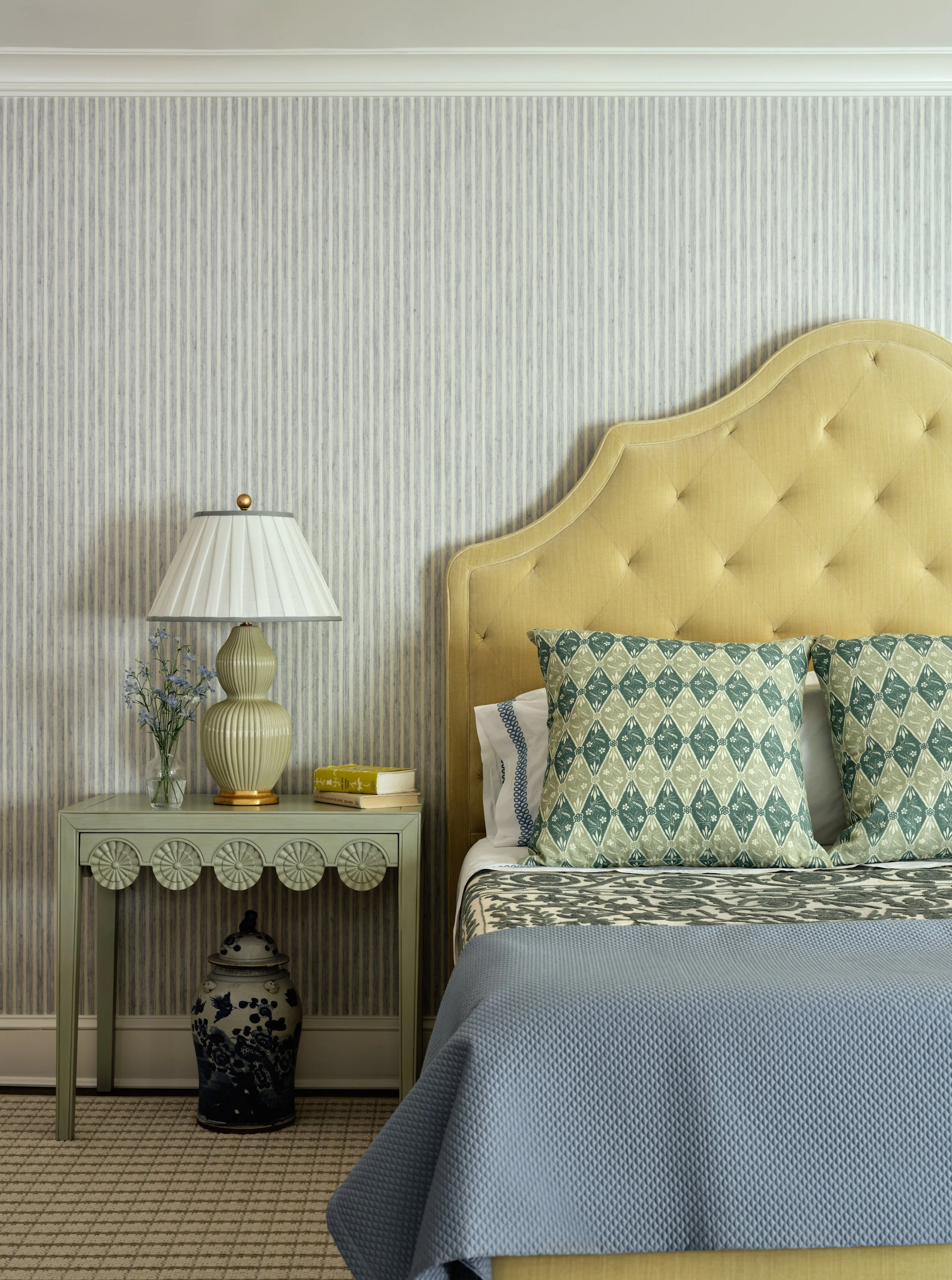 A hotel room with a yellow headboard, patterned pillows, a gray bedspread, a nightstand with a lamp, books, a flower vase, and decorative vase underneath, and striped wallpaper.
