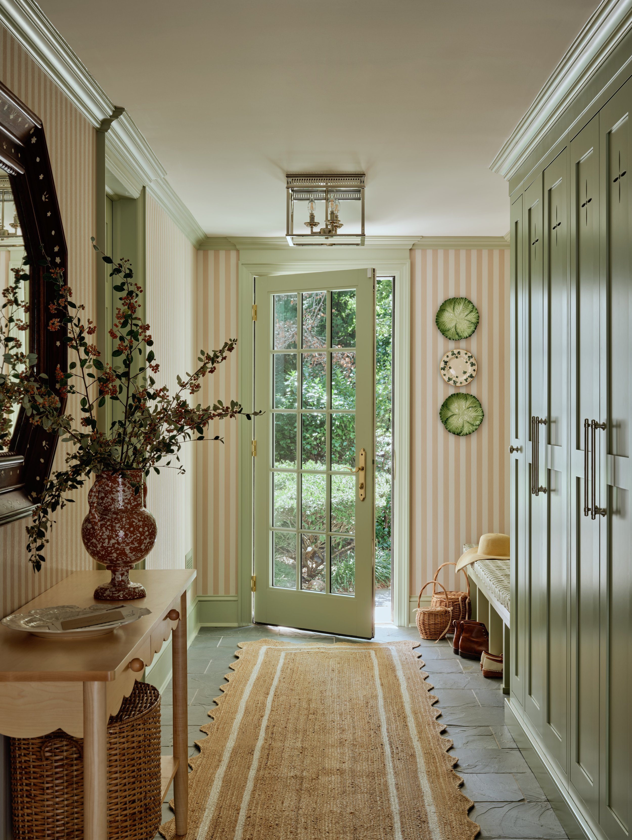 Entryway with a rug, potted plant, and a door opening to a garden, decorated with wall plates, plants, and hallway furniture.