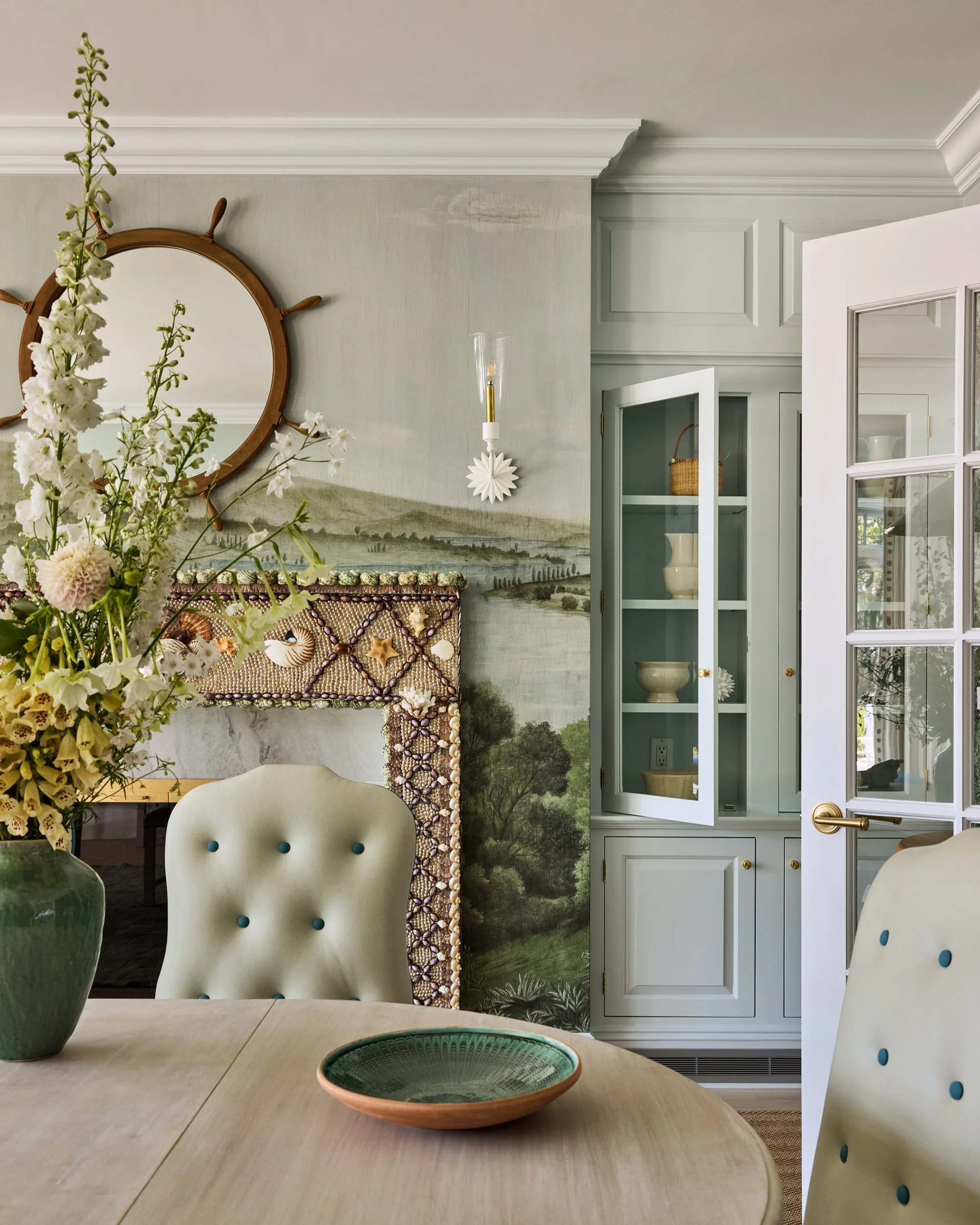 A dining room with a light-colored wooden table, beige upholstered chairs with turquoise buttons, a large green vase with white and light yellow flowers, a decorative mirror resembling a ship's steering wheel, painted landscape mural on the wall, a w