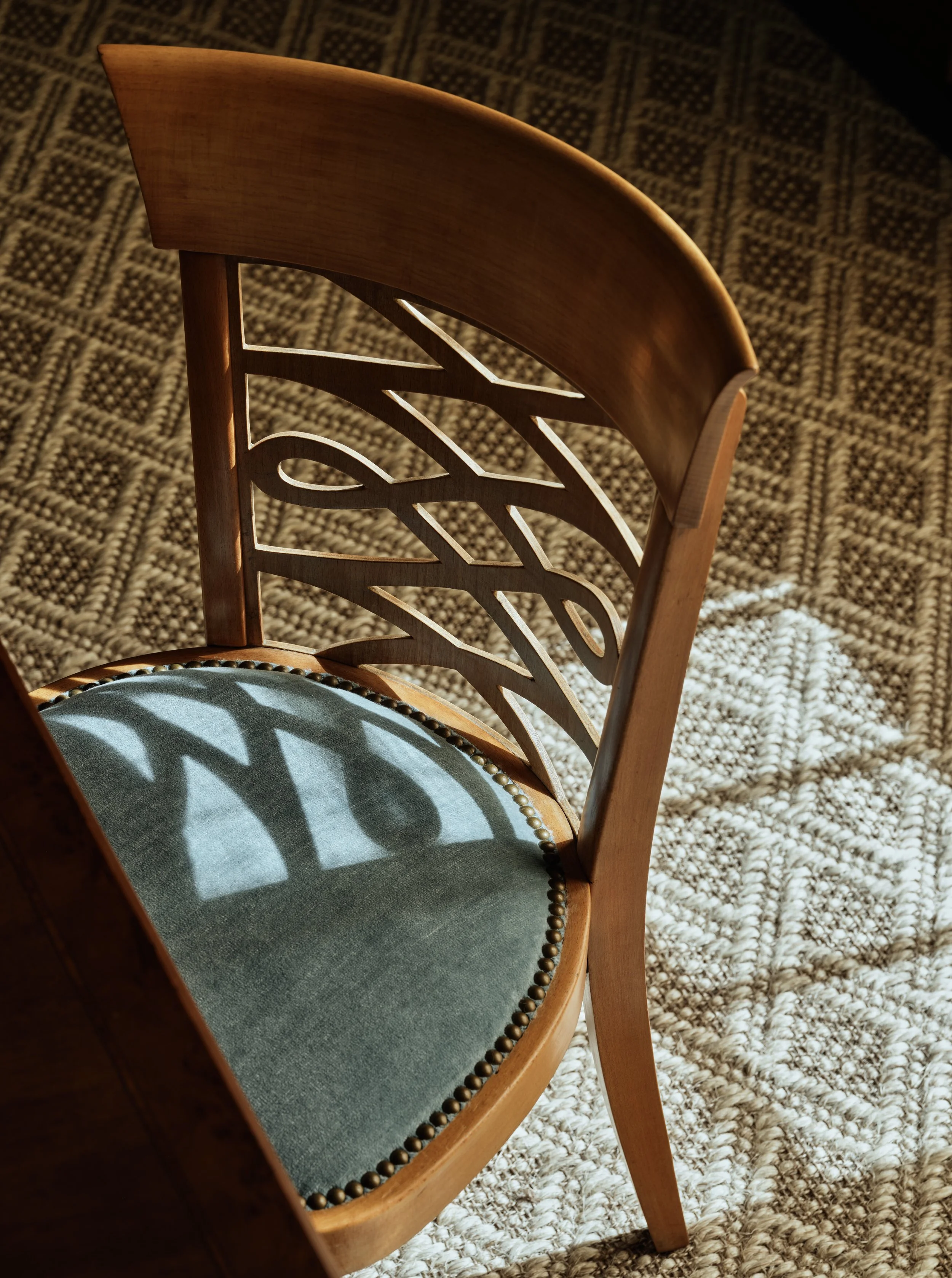 A wooden chair with a decorative backrest and an upholstered seat casts a shadow on a patterned rug.