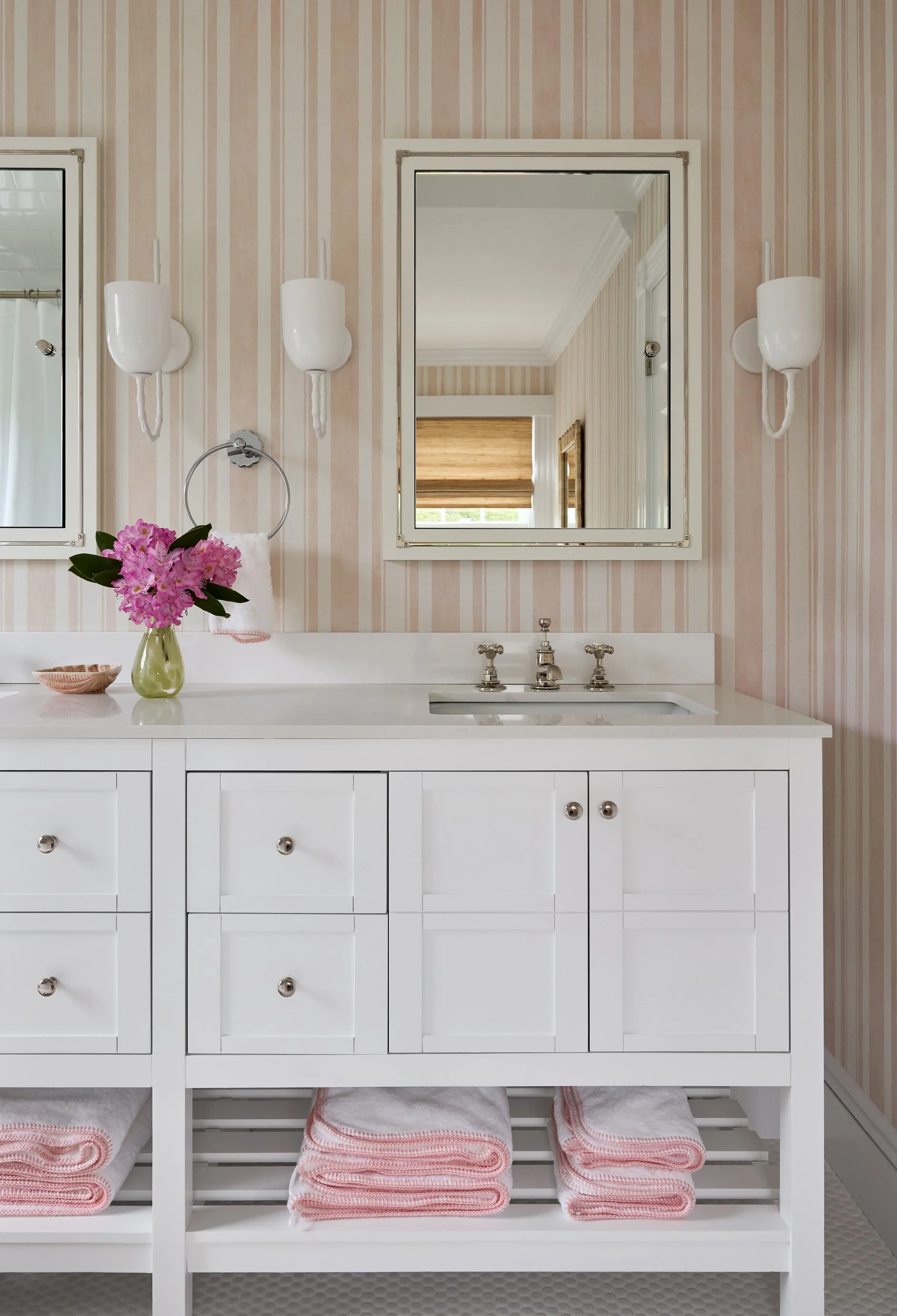 Bathroom vanity with a white cabinet, a mirror, three wall sconces, a pink flower arrangement in a green vase, and pink towels under the sink.