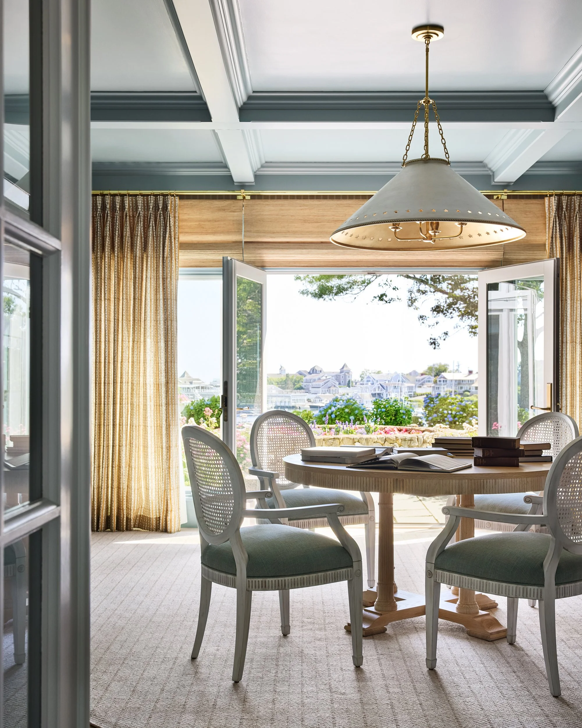 A bright dining room with a round wooden table, four upholstered chairs, and an open set of doors leading to a balcony with a view of houses and trees.