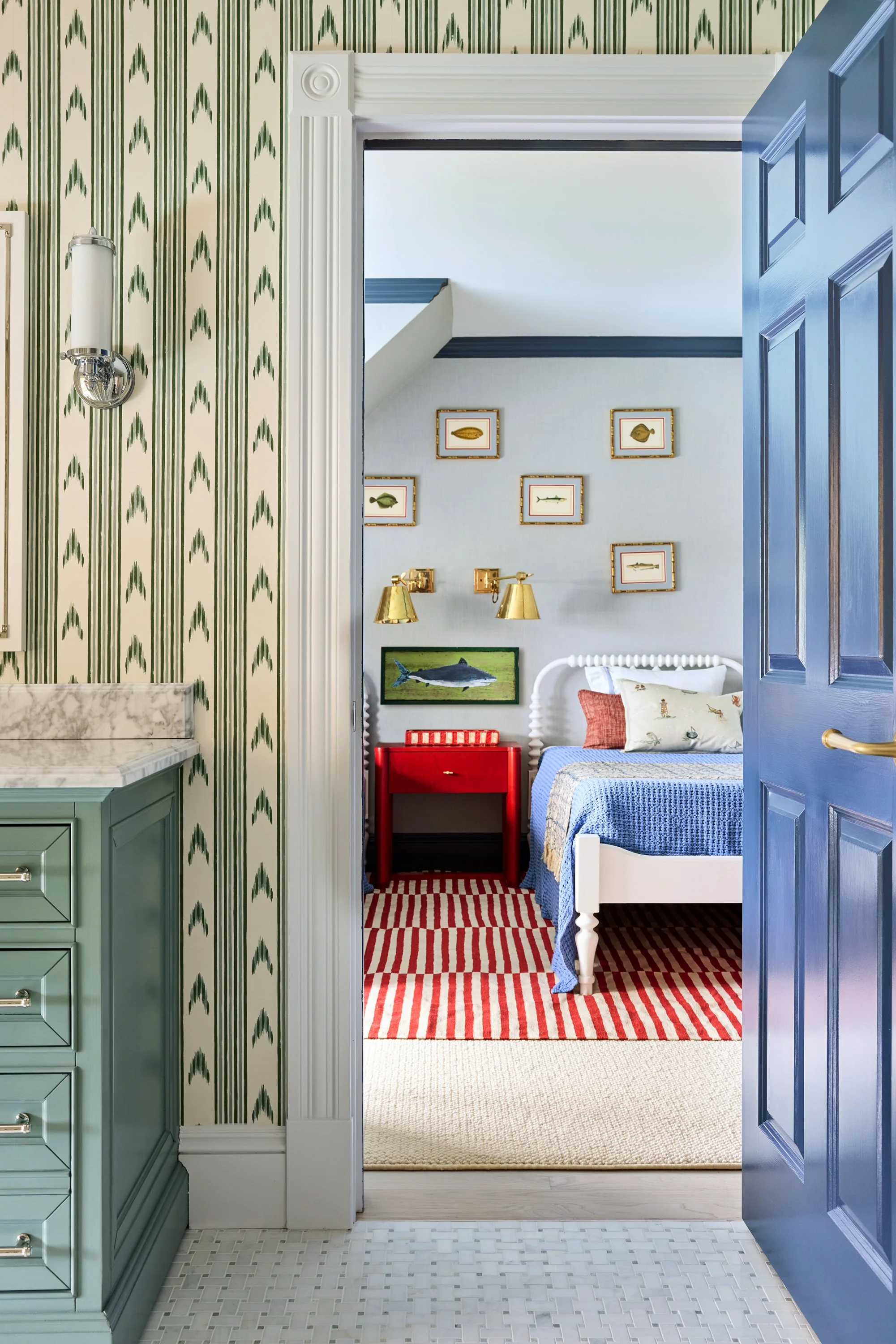 View into a bedroom from a doorway, featuring a white bed with a blue bedspread, a red side table, framed fish artwork on the wall, and a red and white striped rug.
