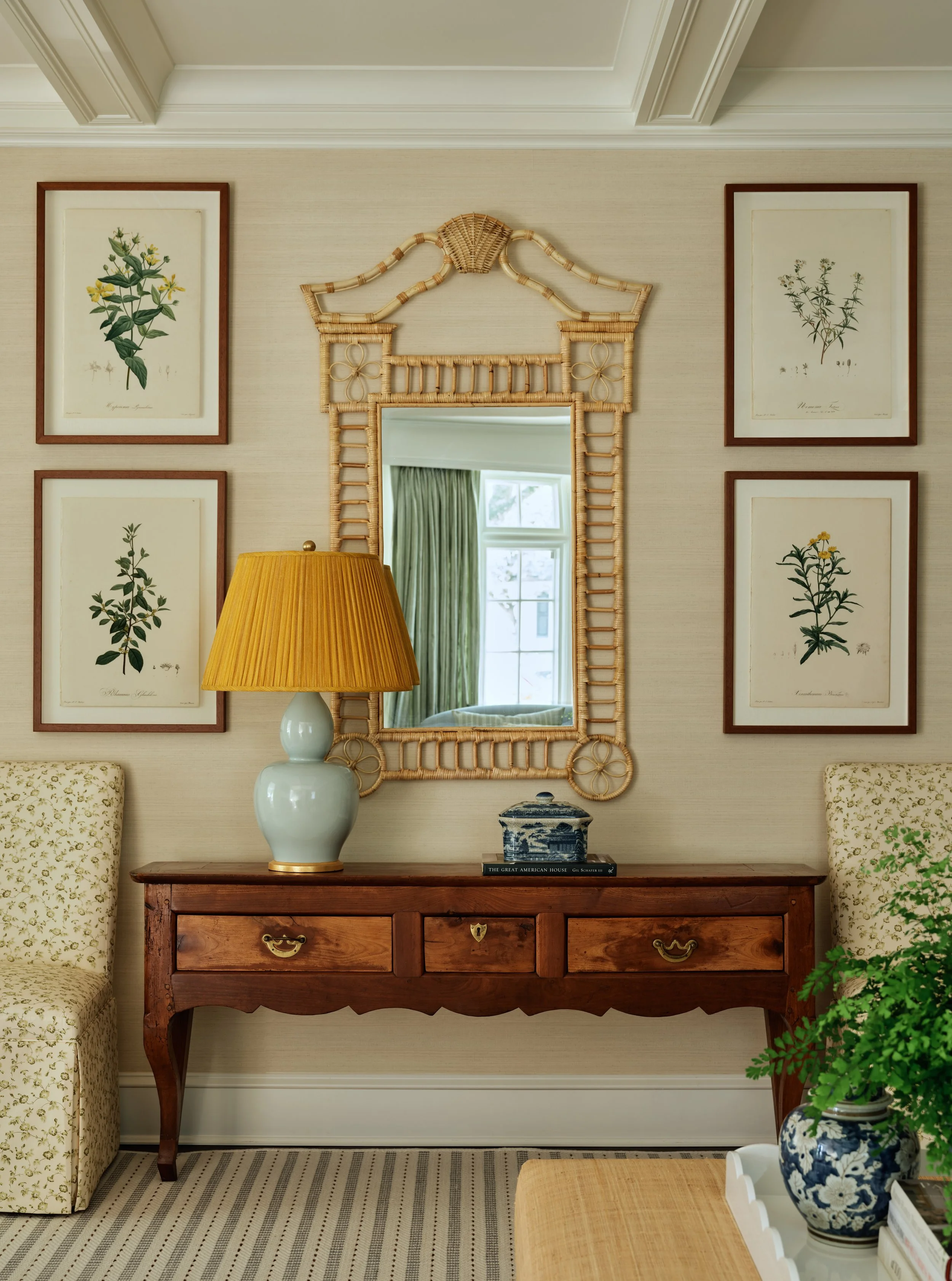 Interior of a living room with a wooden console table, a large yellow lampshade, a decorative mirror framed with wicker, framed botanical prints on the wall, and a reflection of a window with green curtains.