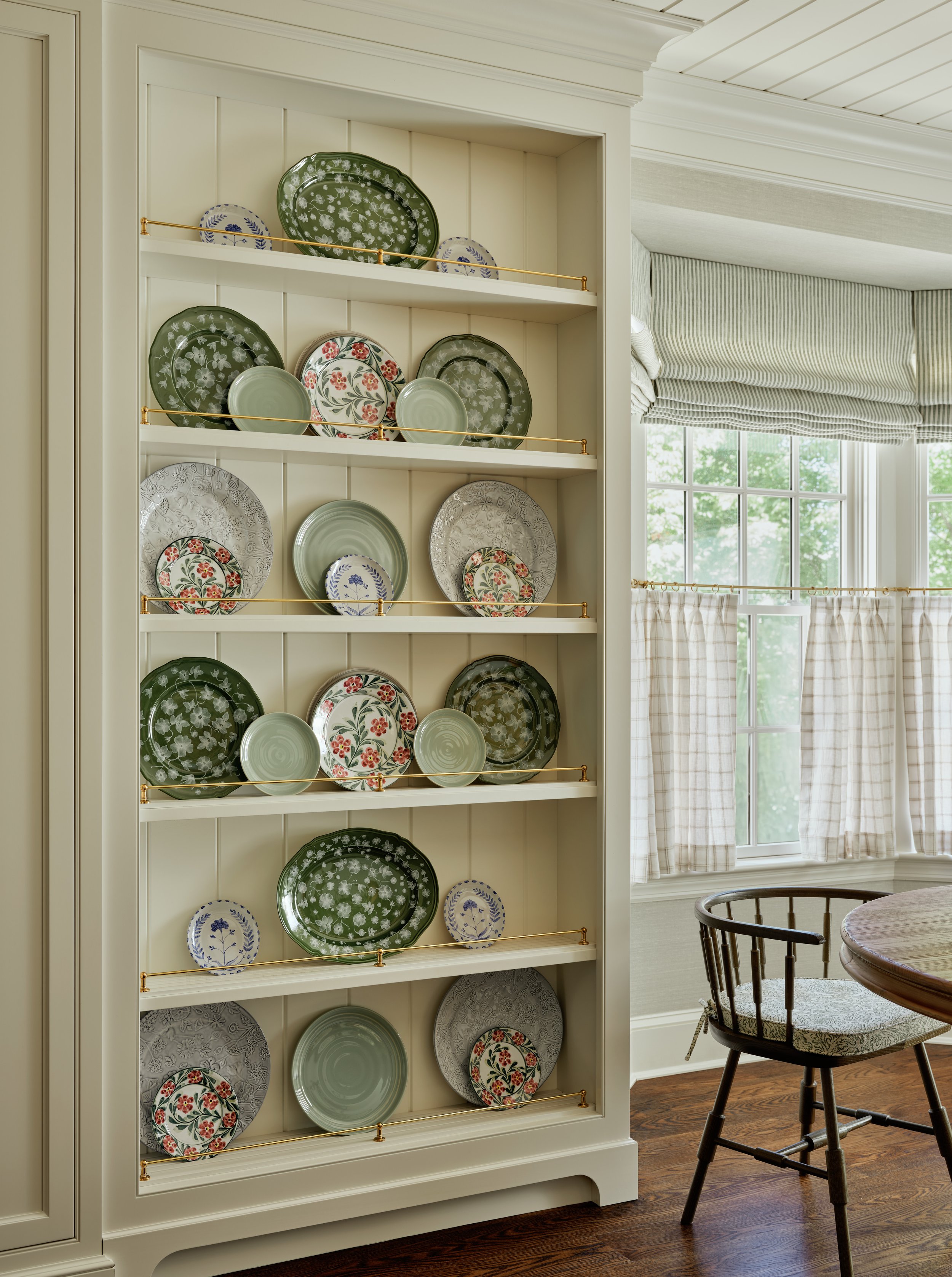 A built-in white cabinet with five shelves holding decorative ceramic plates, some with floral patterns, in a room with wood flooring, large windows, and checkered curtains.